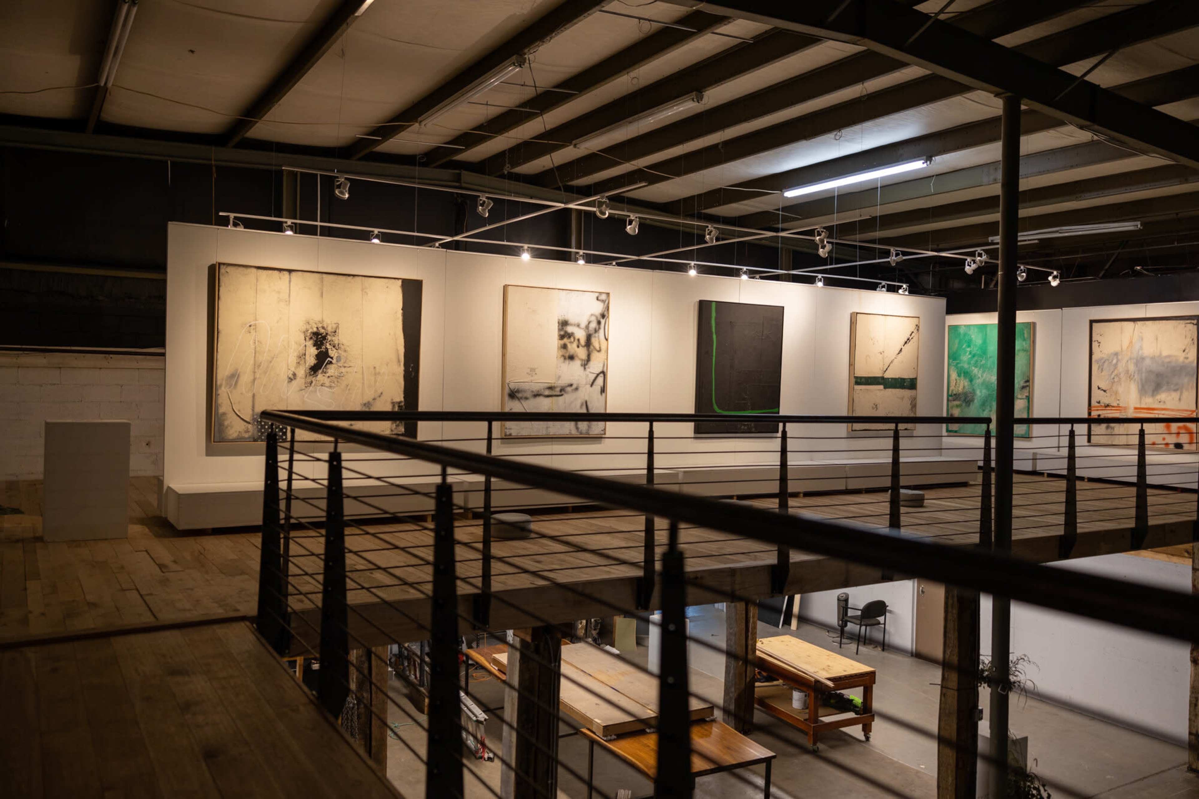 The image shows an art gallery featuring large abstract paintings displayed on white walls, viewed from a balcony overlooking a workshop area.
