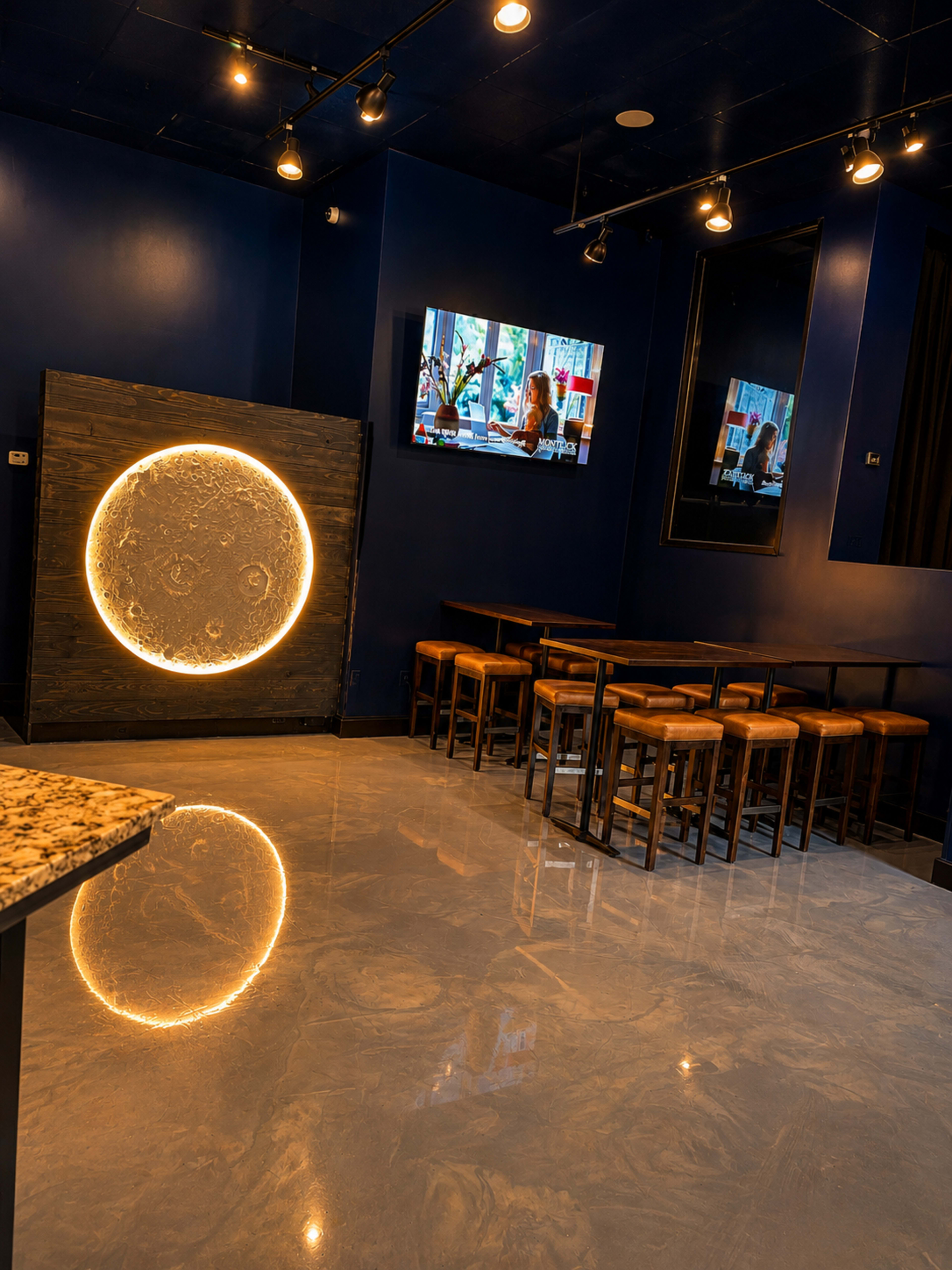 The image shows a modern restaurant interior featuring a dark blue wall with a circular wall design, a television mounted above, and wooden tables with stools arranged in front.
