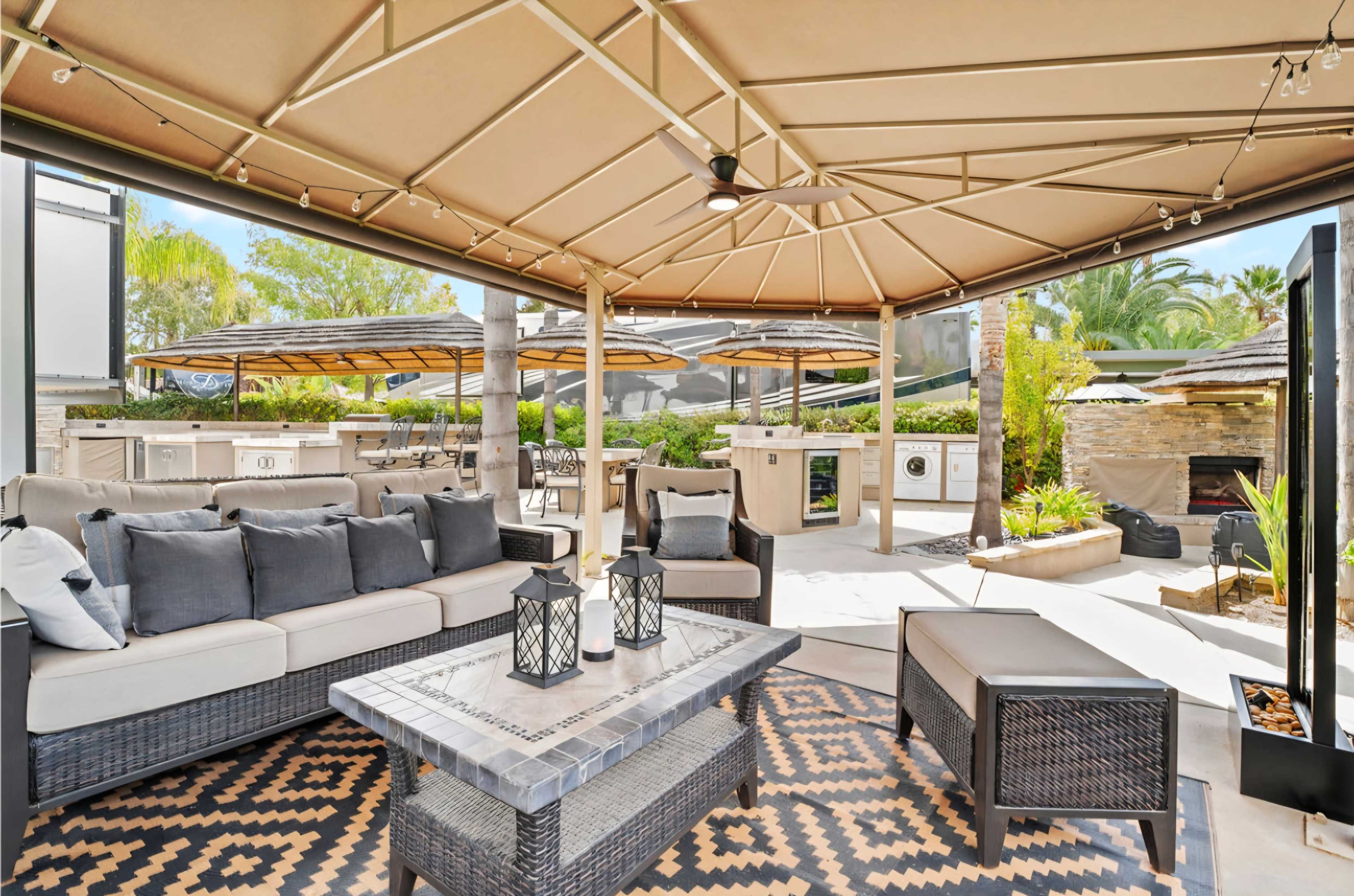 A shaded outdoor seating area features a stylish sofa, coffee table, and lanterns, surrounded by cooking stations and greenery.