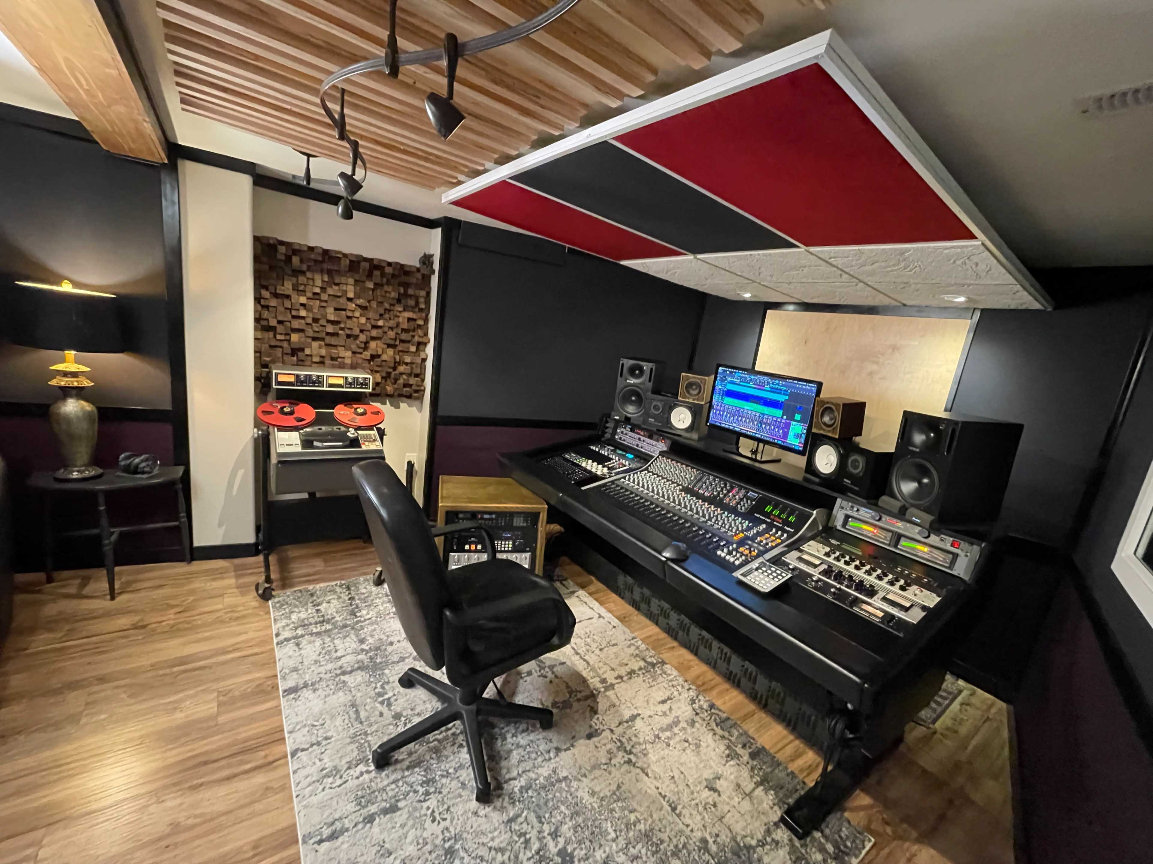 A music studio with a large mixing console, computer monitors, speakers, and soundproof walls, accompanied by a vintage tape deck and an area rug.