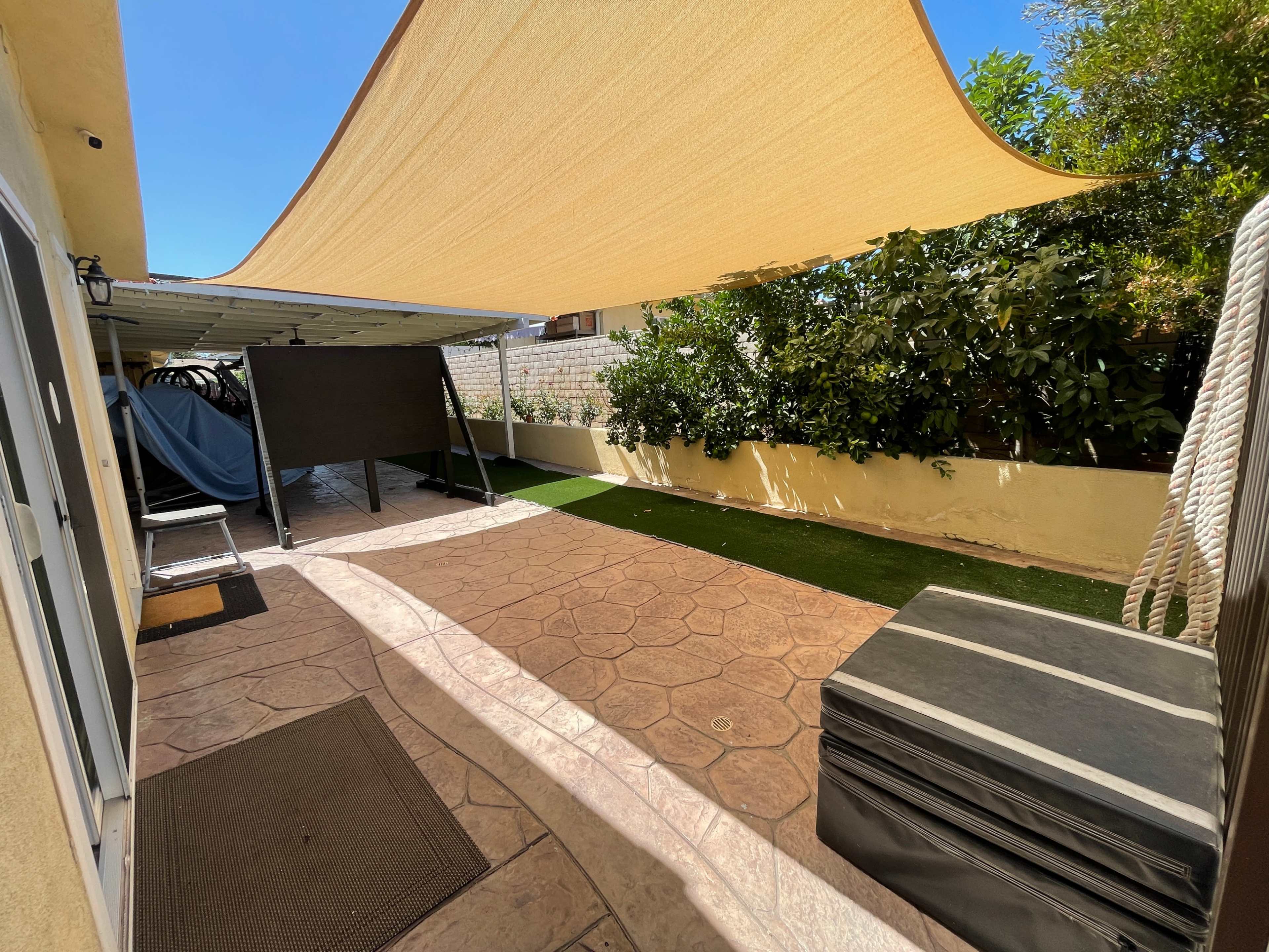 A patio area with a large shade sail overhead, featuring a textured concrete floor, artificial grass, and a covered storage space on one side.