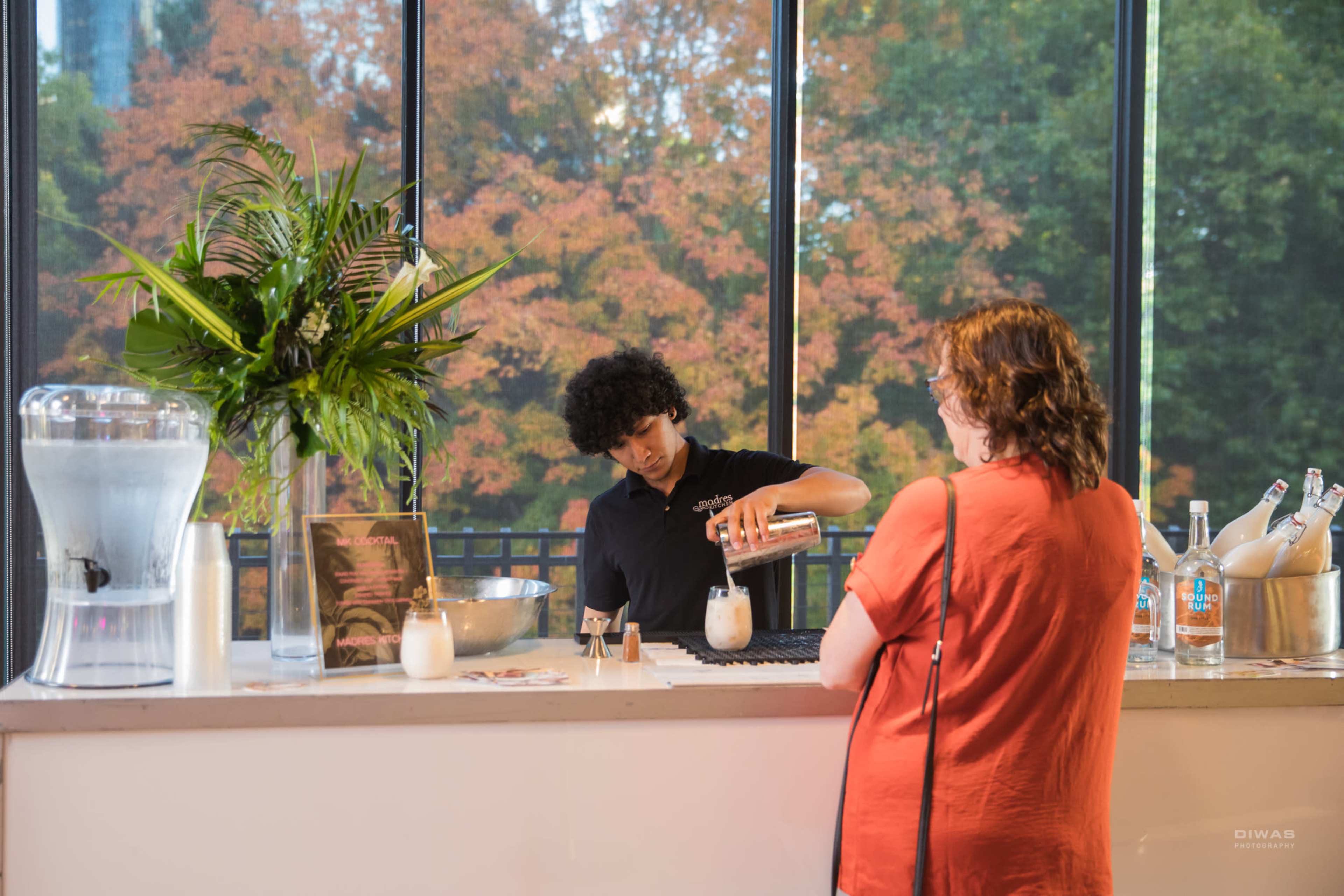A bartender prepares a drink for a customer at a bar with large windows showing trees in autumn colors outside.