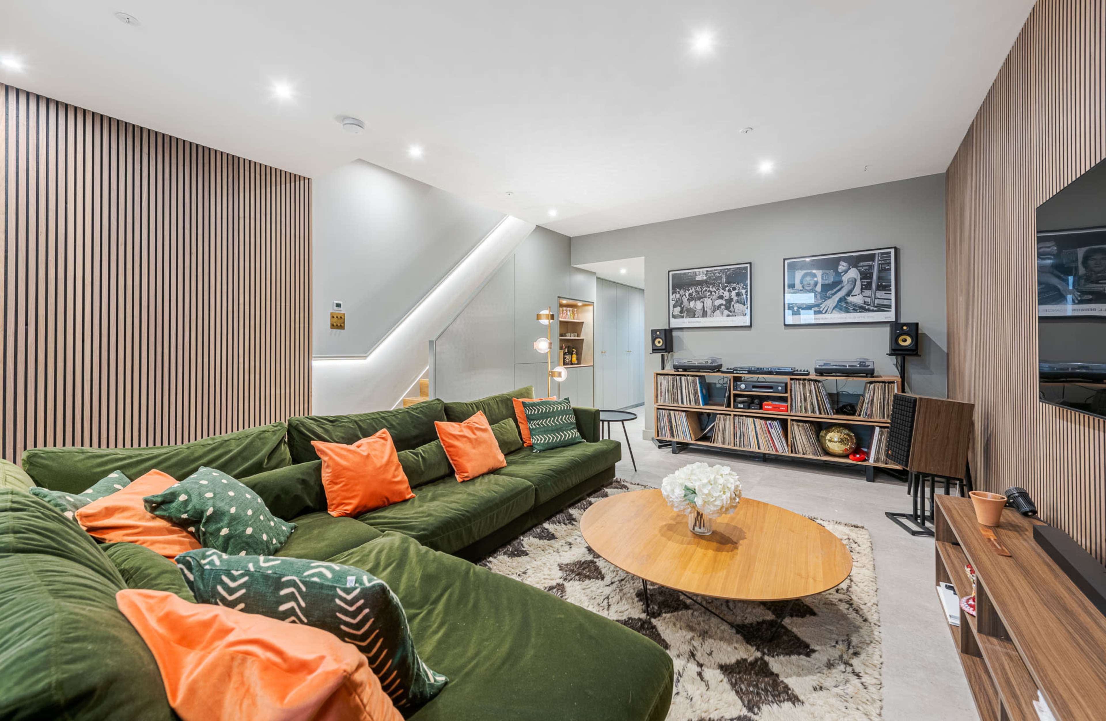 The living room features a green sofa with orange cushions, a round wooden coffee table, and framed photographs on the walls, alongside shelves containing vinyl records.