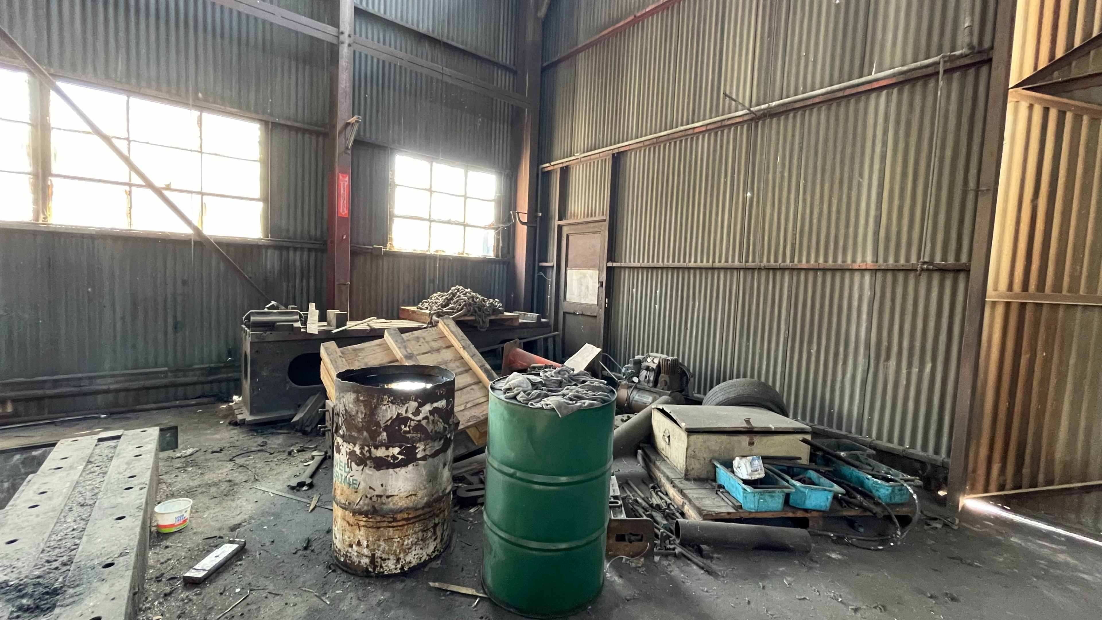 The image shows a cluttered industrial space with various discarded items, including barrels, wooden pallets, and debris scattered across the floor.