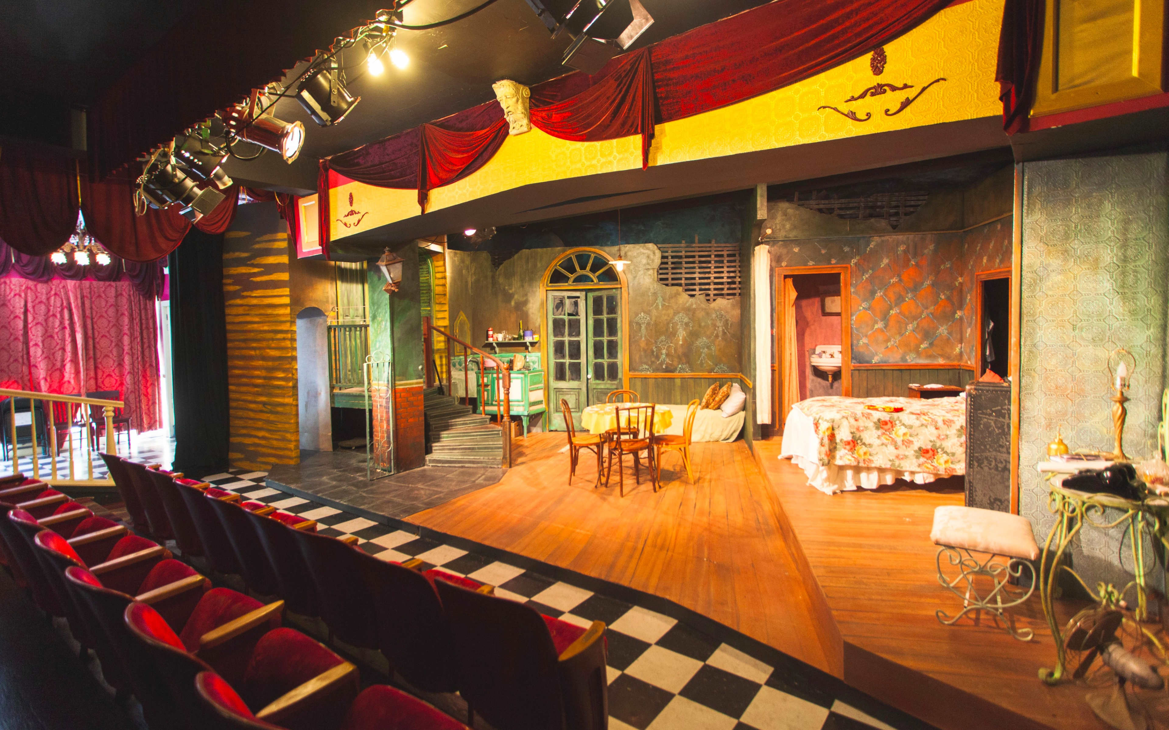 A theater stage set with a colorful backdrop, furniture, and stage lighting, viewed from the audience seating area.