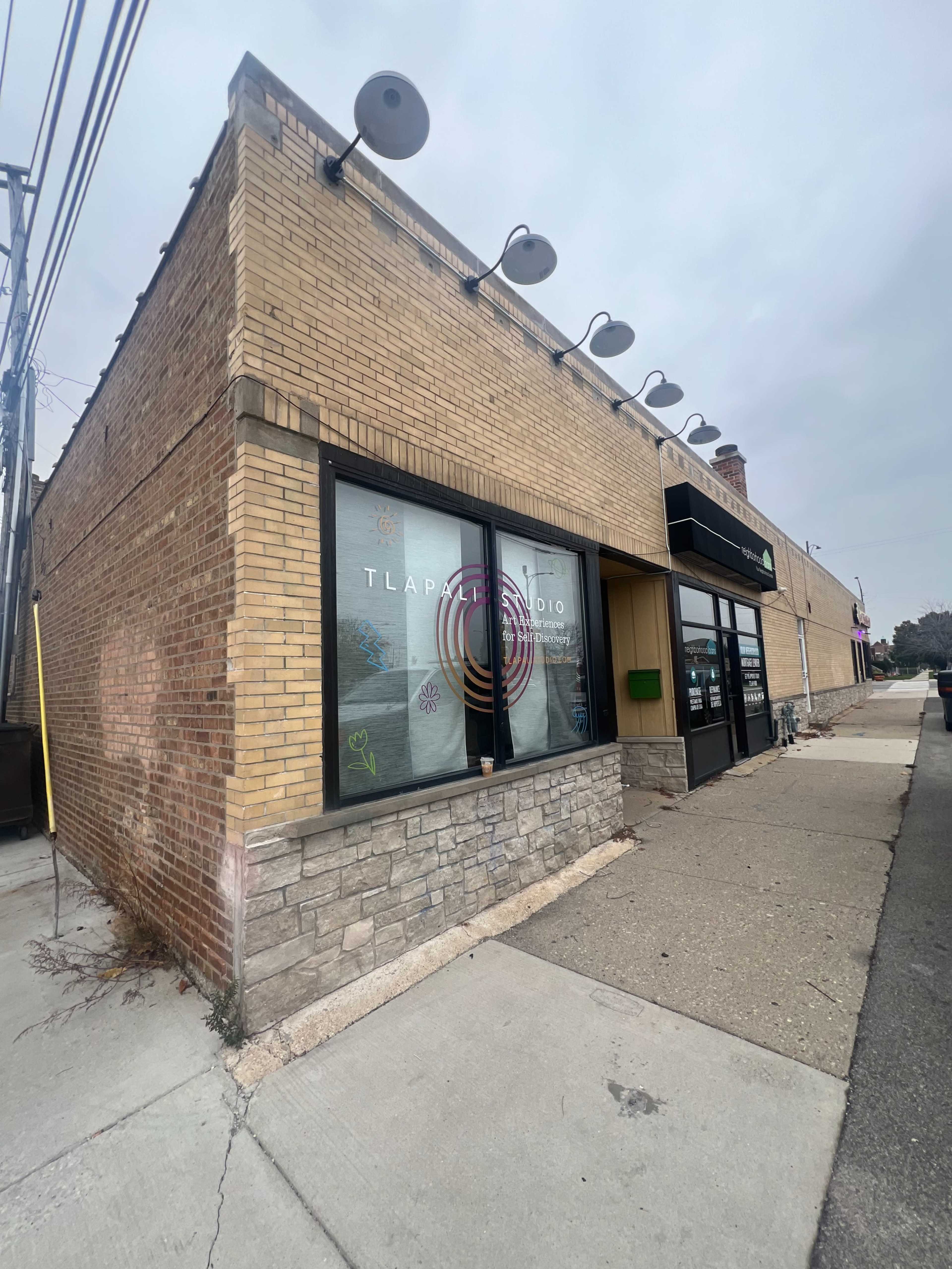 The image shows a corner commercial building with a brick exterior, large windows featuring colorful designs, and a sidewalk in front.