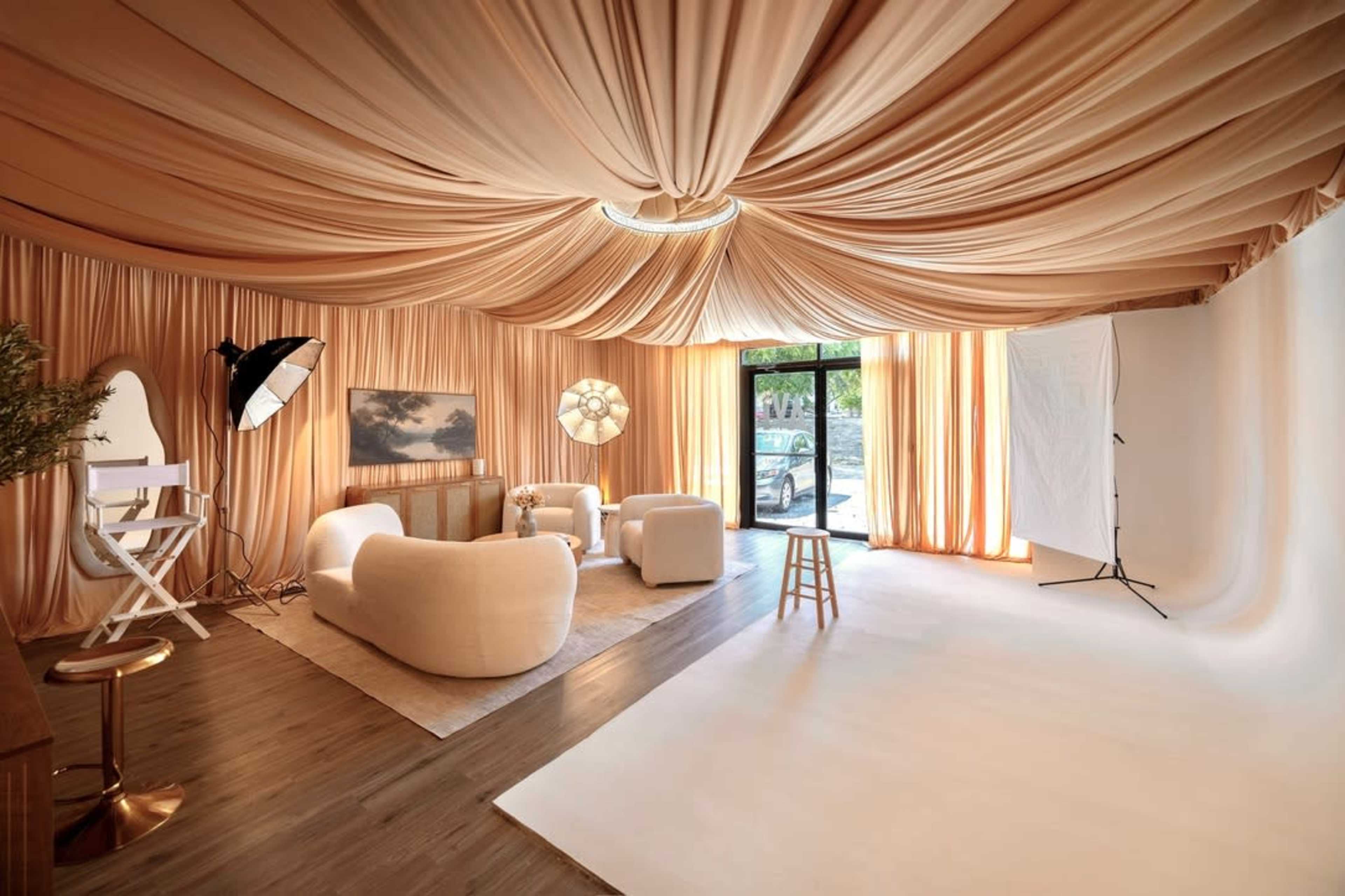 The image shows a well-lit studio space with draped beige fabric covering the walls and ceiling, featuring modern furniture, soft lighting, and a large window.
