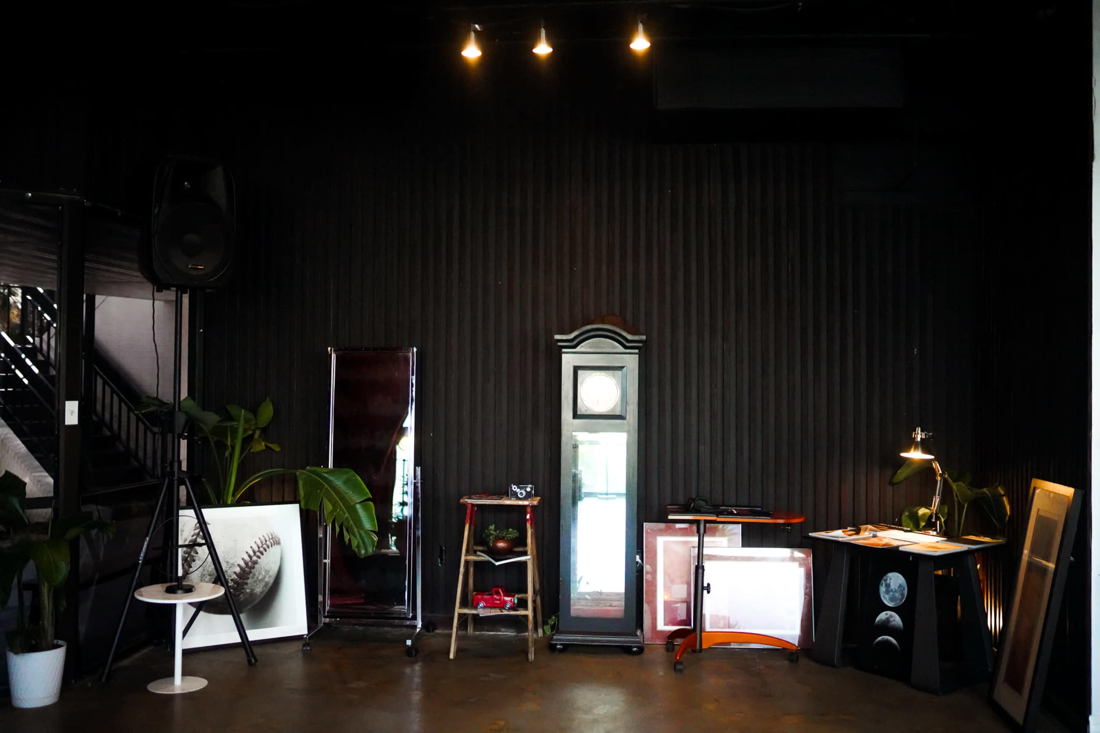 A dark industrial space features various mirrors, a small table, plants, and a standing lamp.