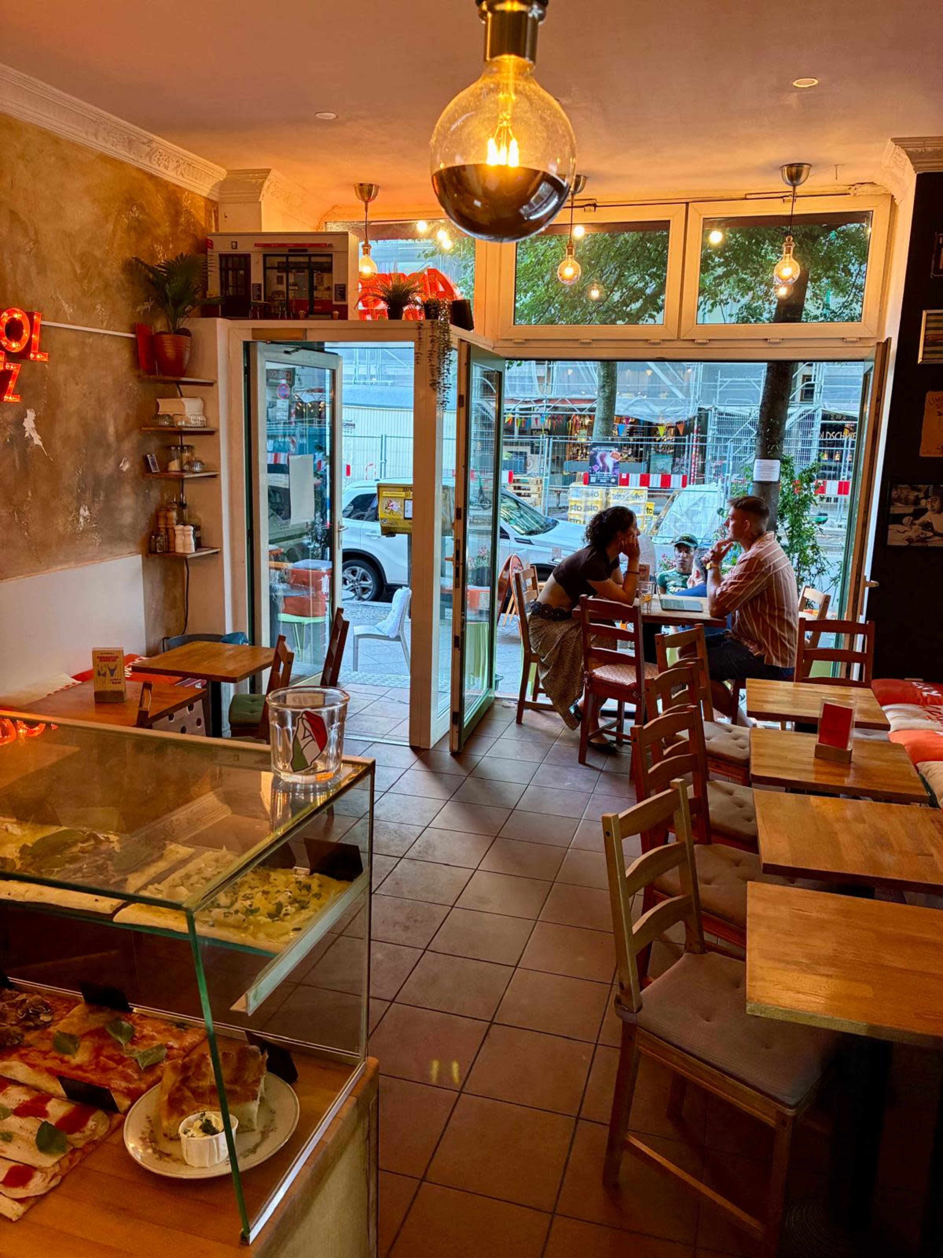 The image shows the interior of a small café with a glass display case filled with food, wooden tables and chairs, and patrons seated near the entrance.