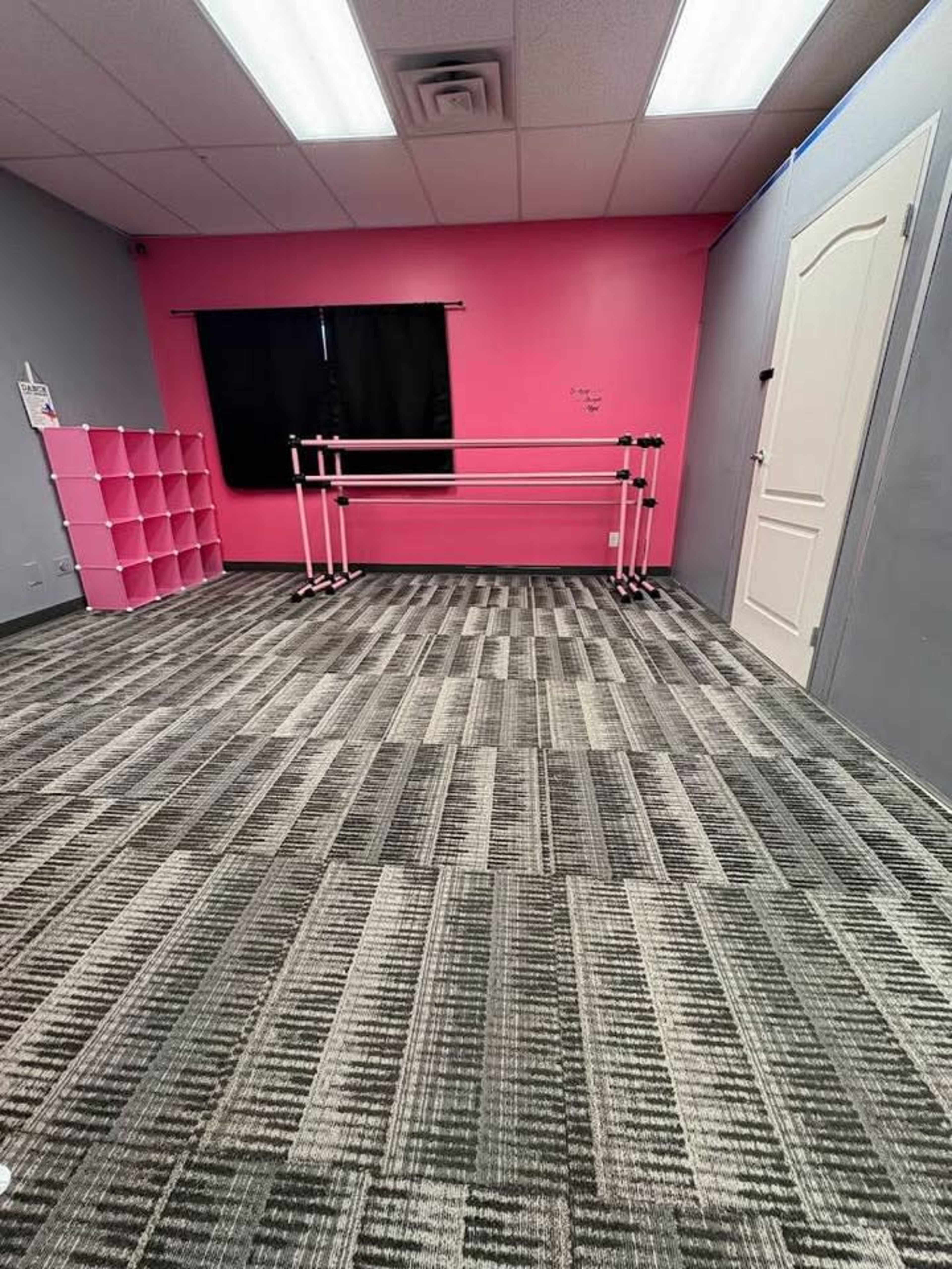 The image shows a spacious dance studio with gray walls, a pink accent wall, a set of ballet bars, and a storage shelf.