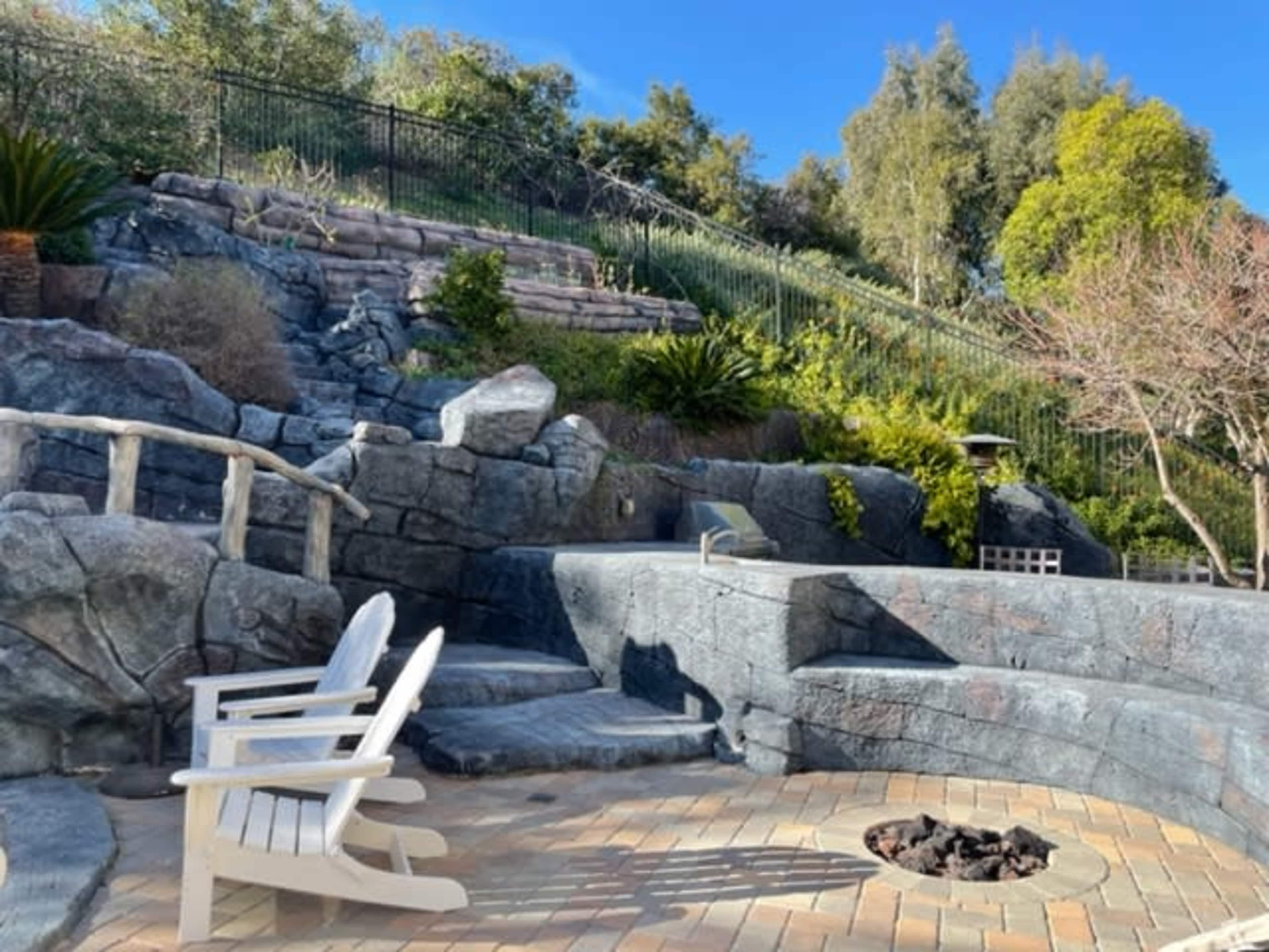 The image shows a landscaped outdoor area featuring stone seating, a fire pit, and lush greenery against a hillside backdrop.