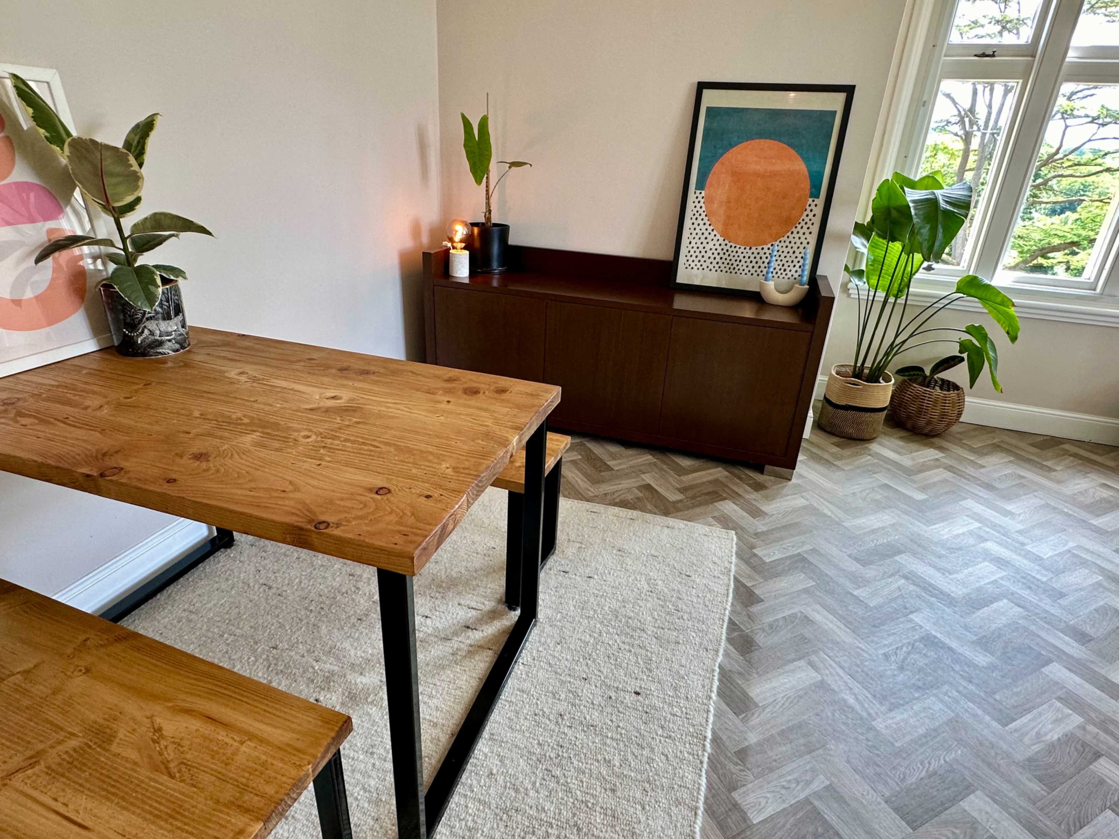 A wooden dining table and benches are positioned on a herringbone-patterned floor beside a sideboard adorned with a framed artwork and potted plants.