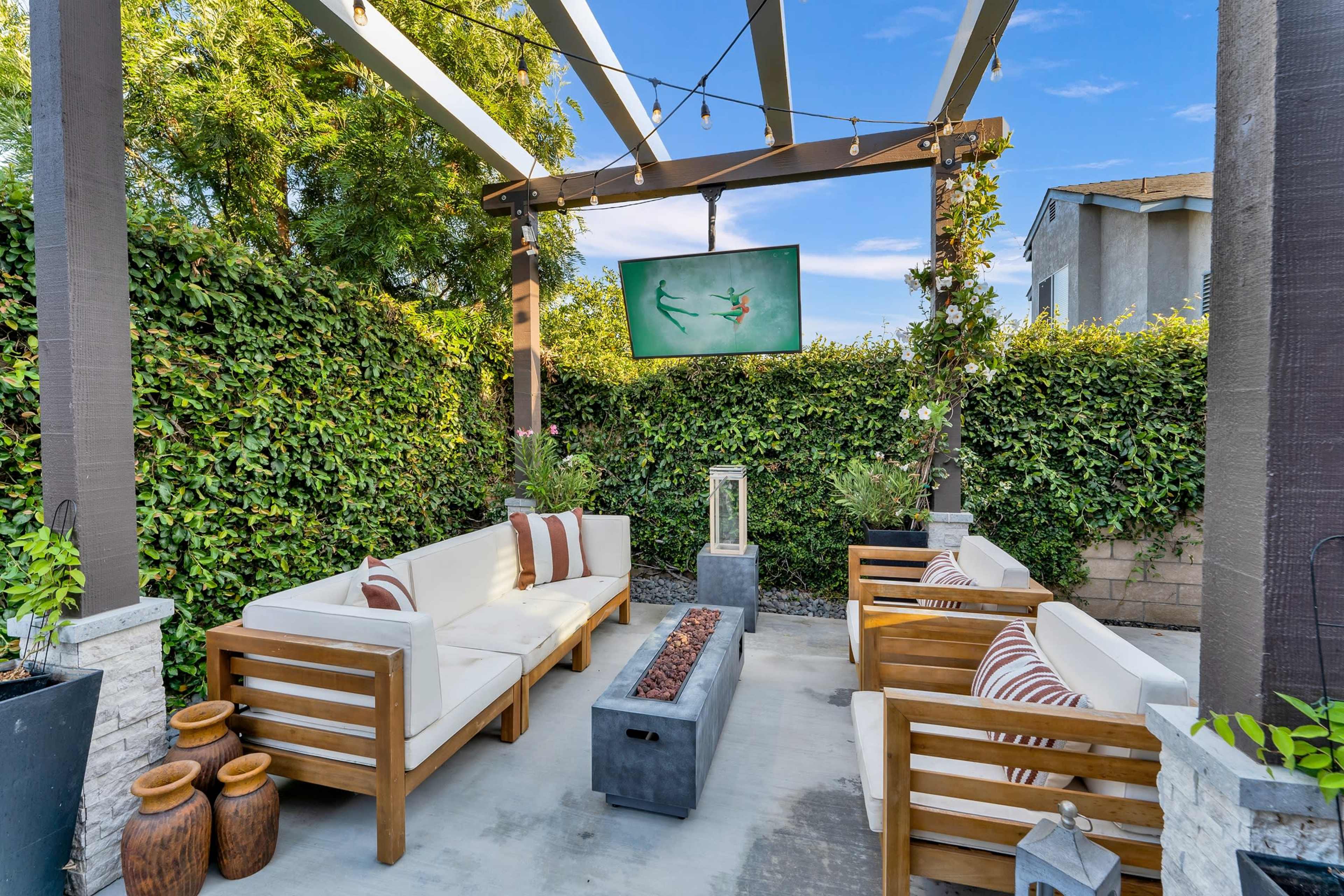 An outdoor seating area with wooden furniture, a flat-screen TV mounted above, and greenery surrounding the space.