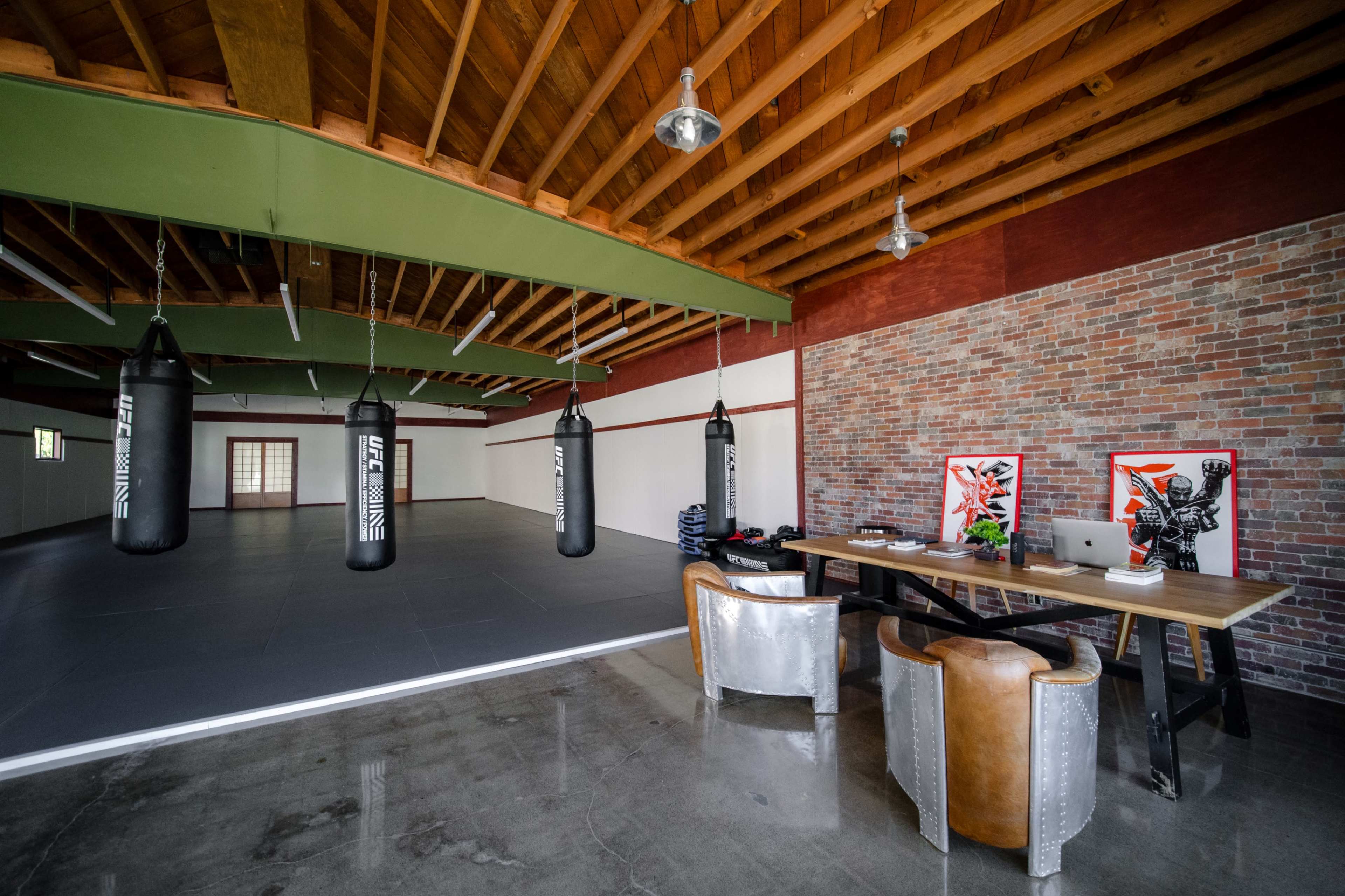 The image shows a spacious training area with four hanging punching bags and a wooden table with chairs set up against a brick wall.