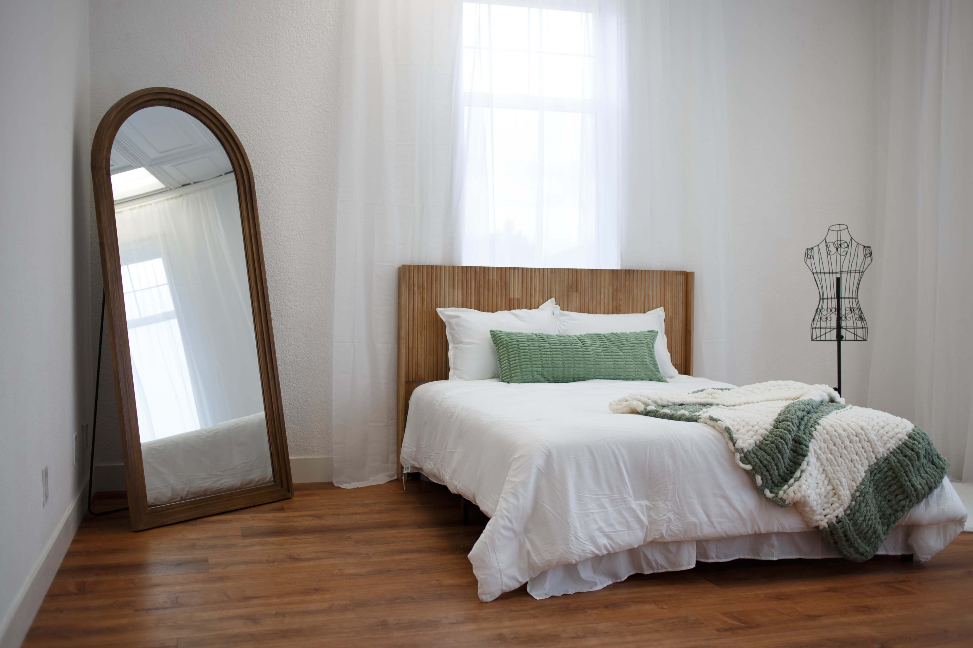 The image shows a neatly arranged bedroom featuring a bed with a wooden headboard, a large mirror, and a dressmaker's form.