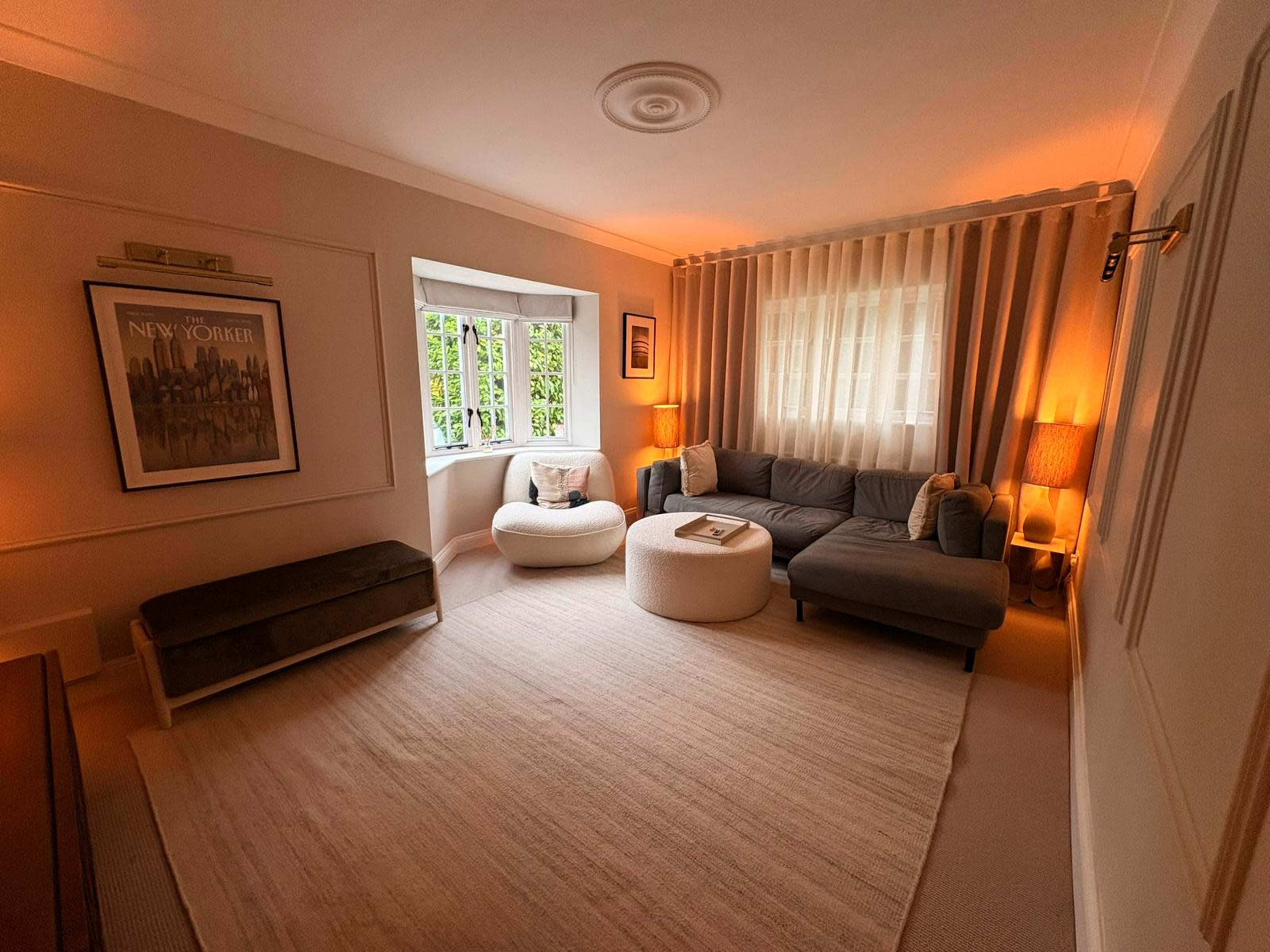 The image shows a cozy living room with a gray sofa, a round coffee table, a plush rug, and soft lighting from lamps and windows.