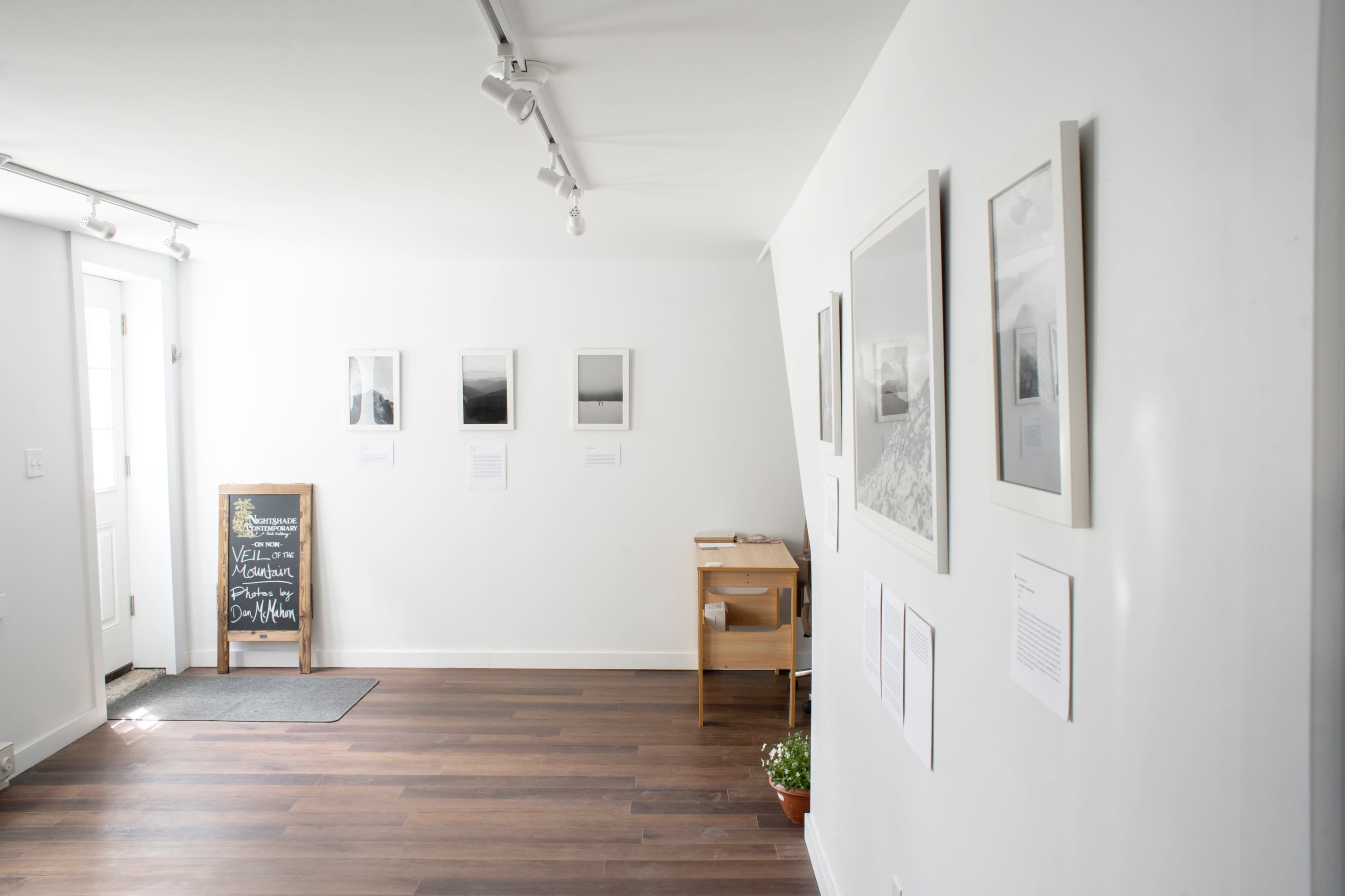 A minimalist gallery space with framed black-and-white photographs on the wall, a wooden table, and a chalkboard near the entrance.