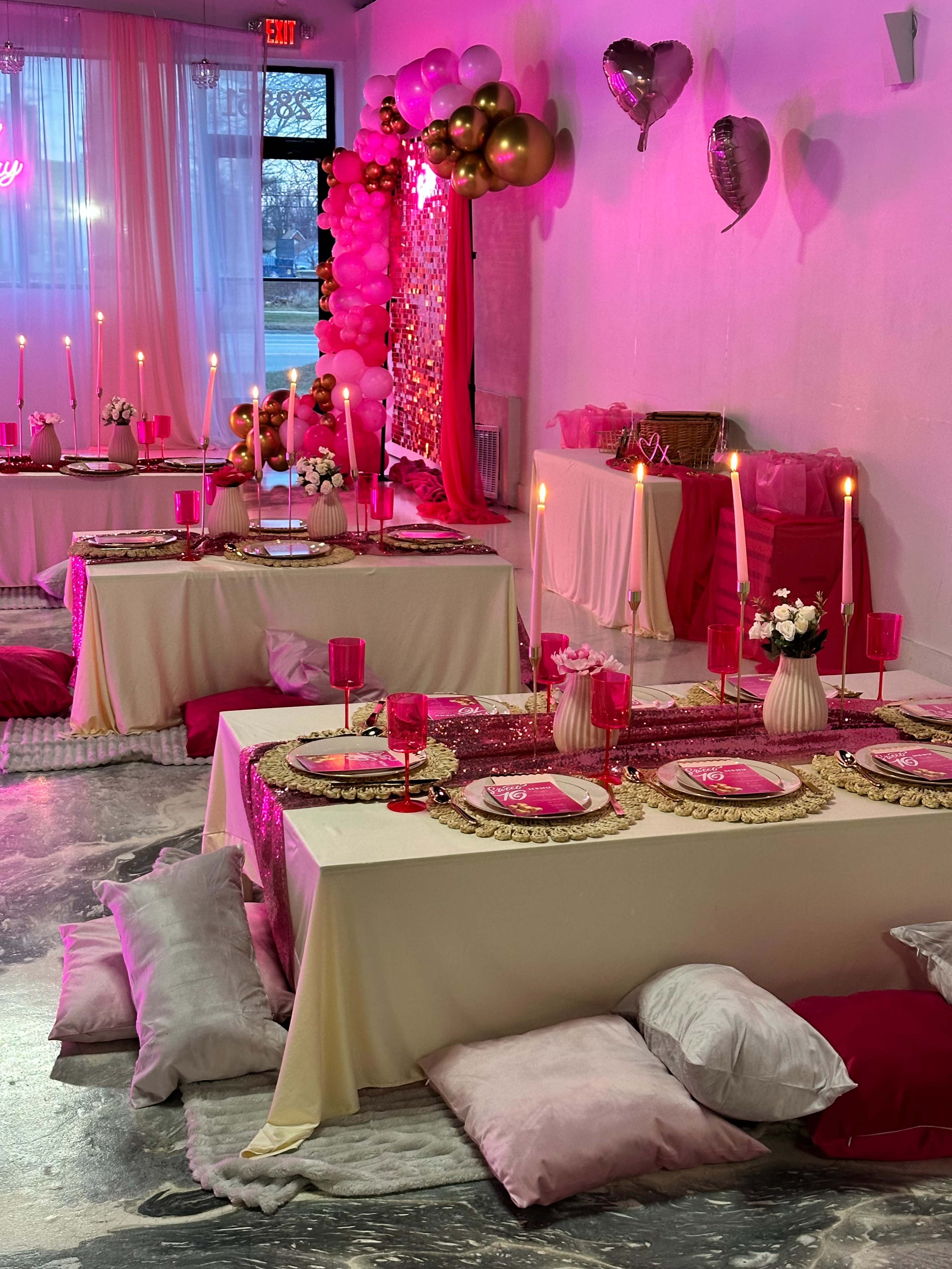 The image shows a decorated event space with tables set for a celebration, featuring pink and gold décor, candles, and plush pillows scattered on the floor.