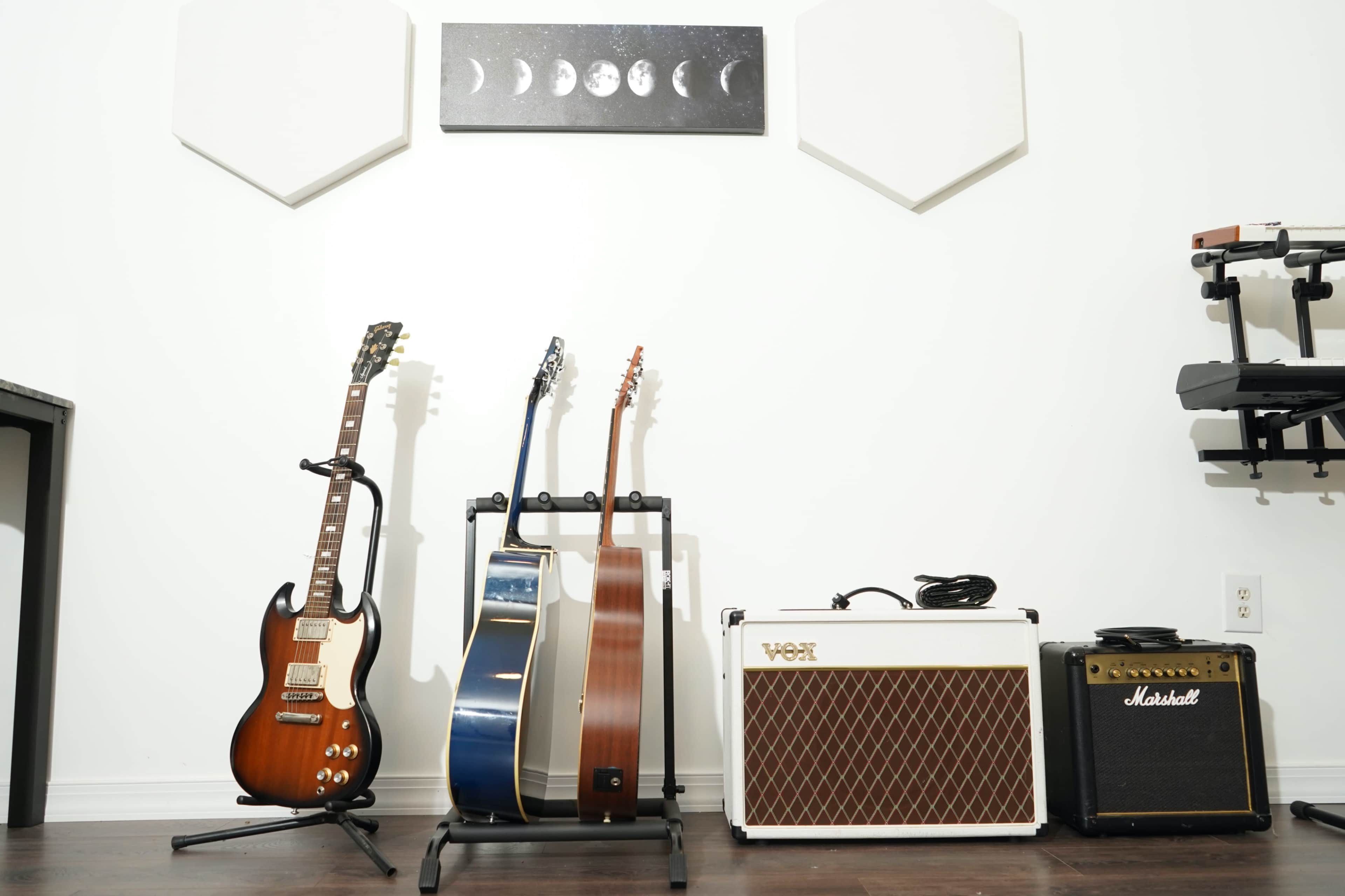 A collection of guitars resting on a stand and amplifiers positioned against a white wall featuring a lunar phases artwork.