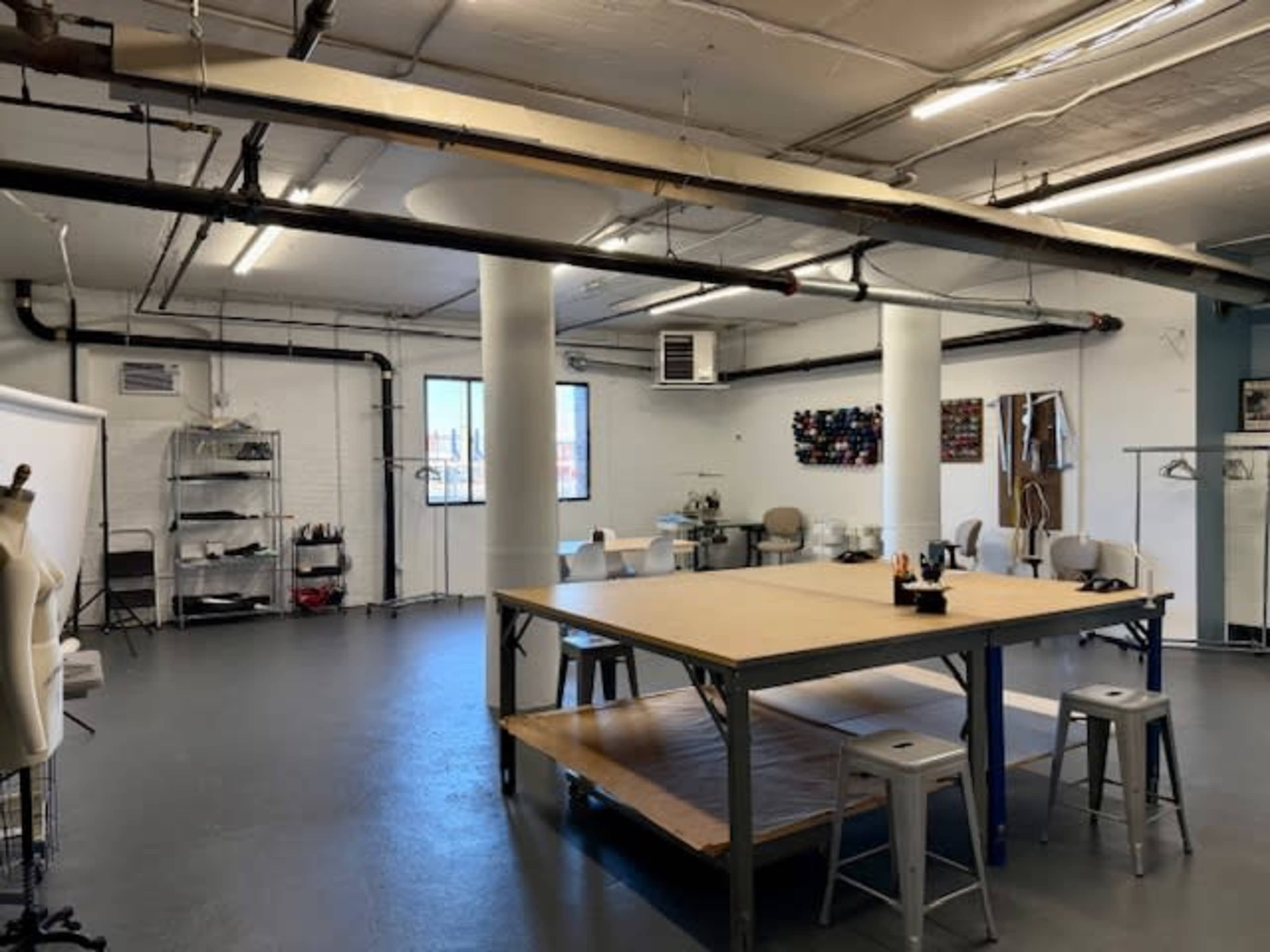 The image shows a spacious studio with large tables, chairs, and storage units, featuring a clean, industrial design with exposed pipes and ample natural light.