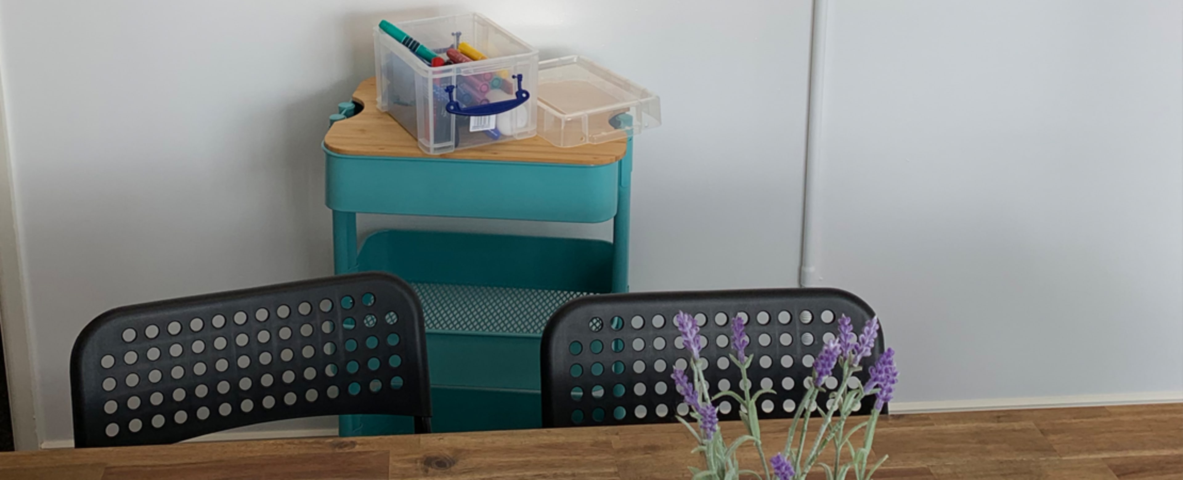 A table with a potted plant sits in front of three black chairs, alongside a teal storage unit holding art supplies.