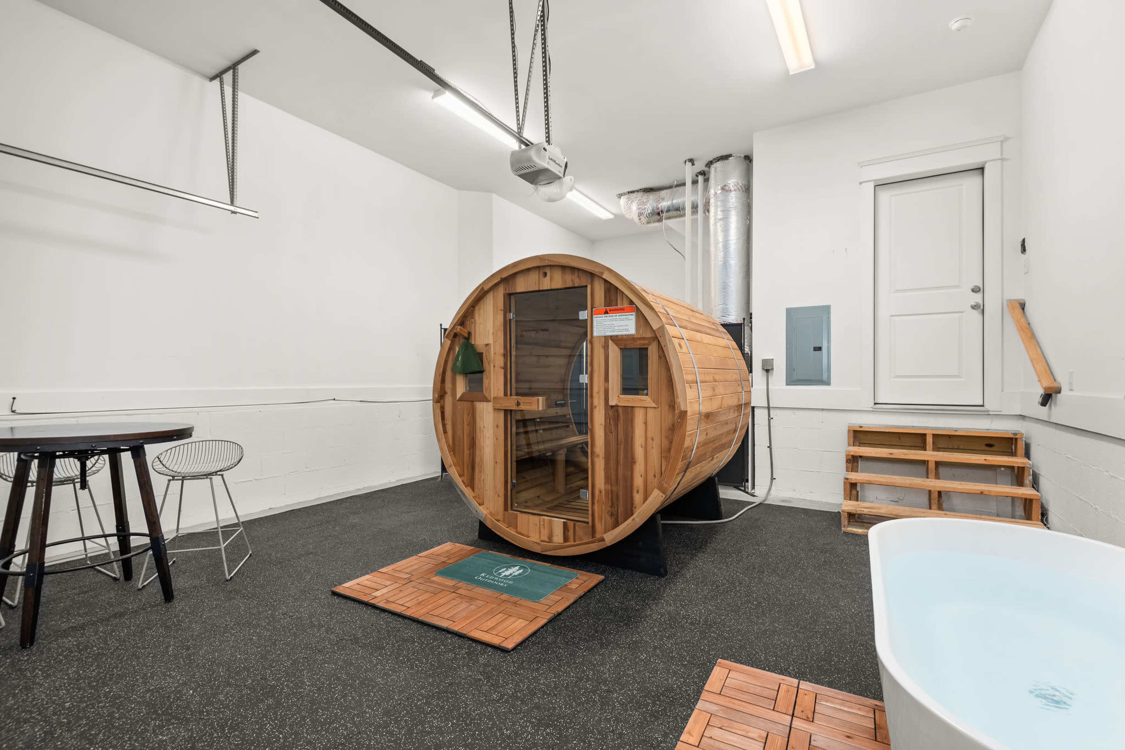 The image shows a wooden sauna in a modern room with gray flooring, a bathtub, and minimalist furnishings.