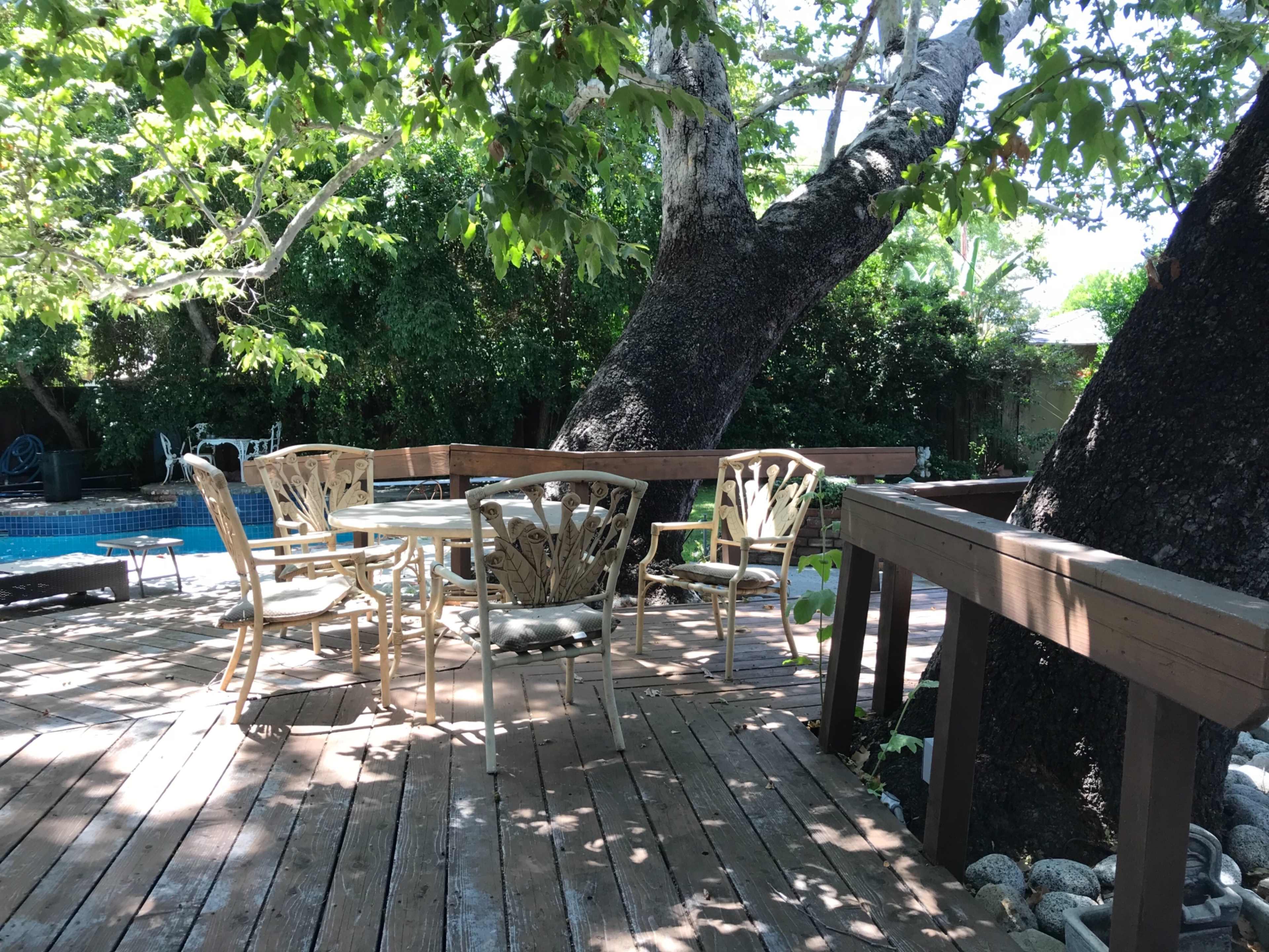 A wooden deck features white outdoor furniture surrounded by trees, with a swimming pool visible in the background.