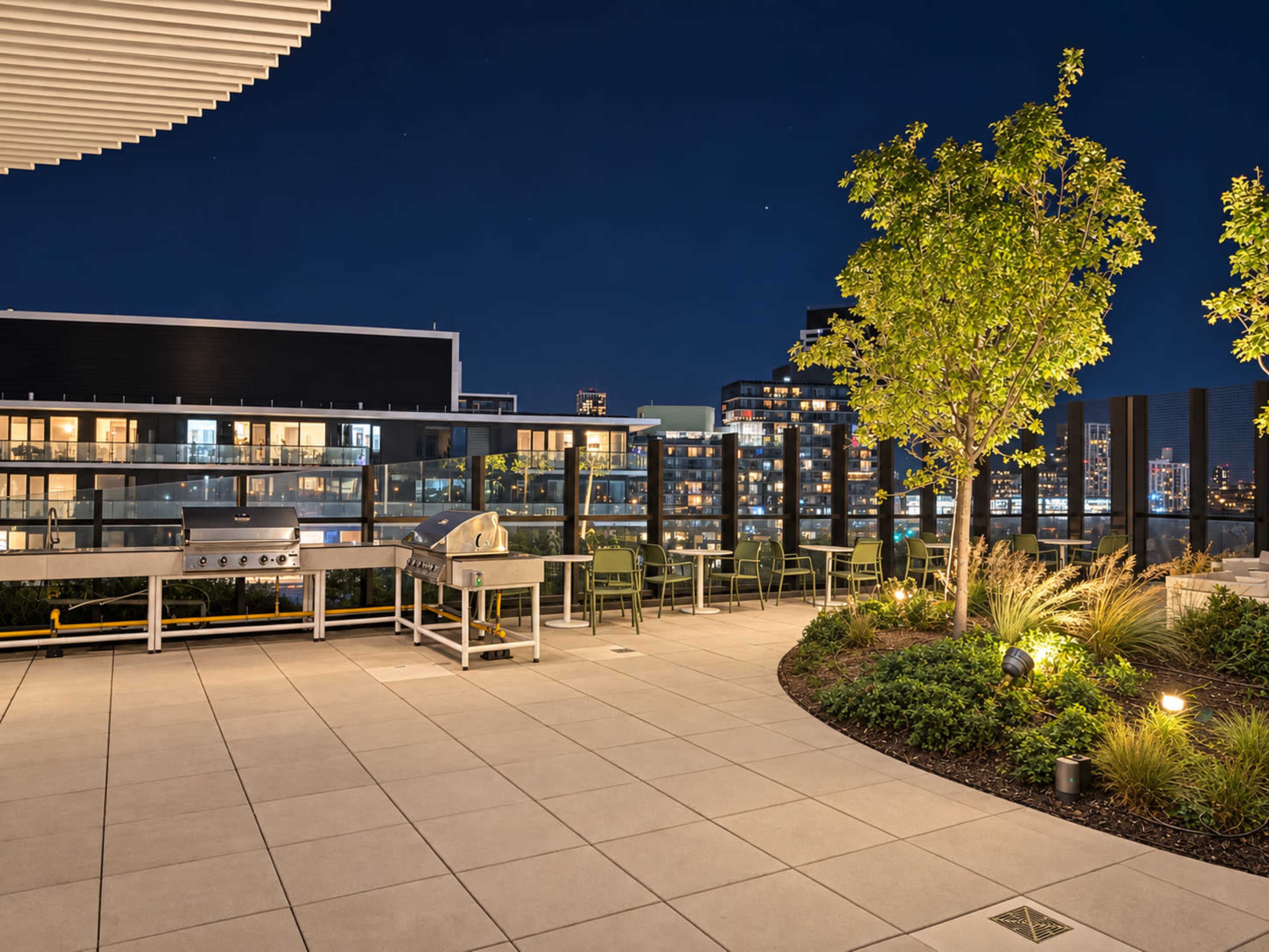 The image shows a modern rooftop terrace at night, featuring a barbecue grill, green seating, and landscaped plants with city lights in the background.