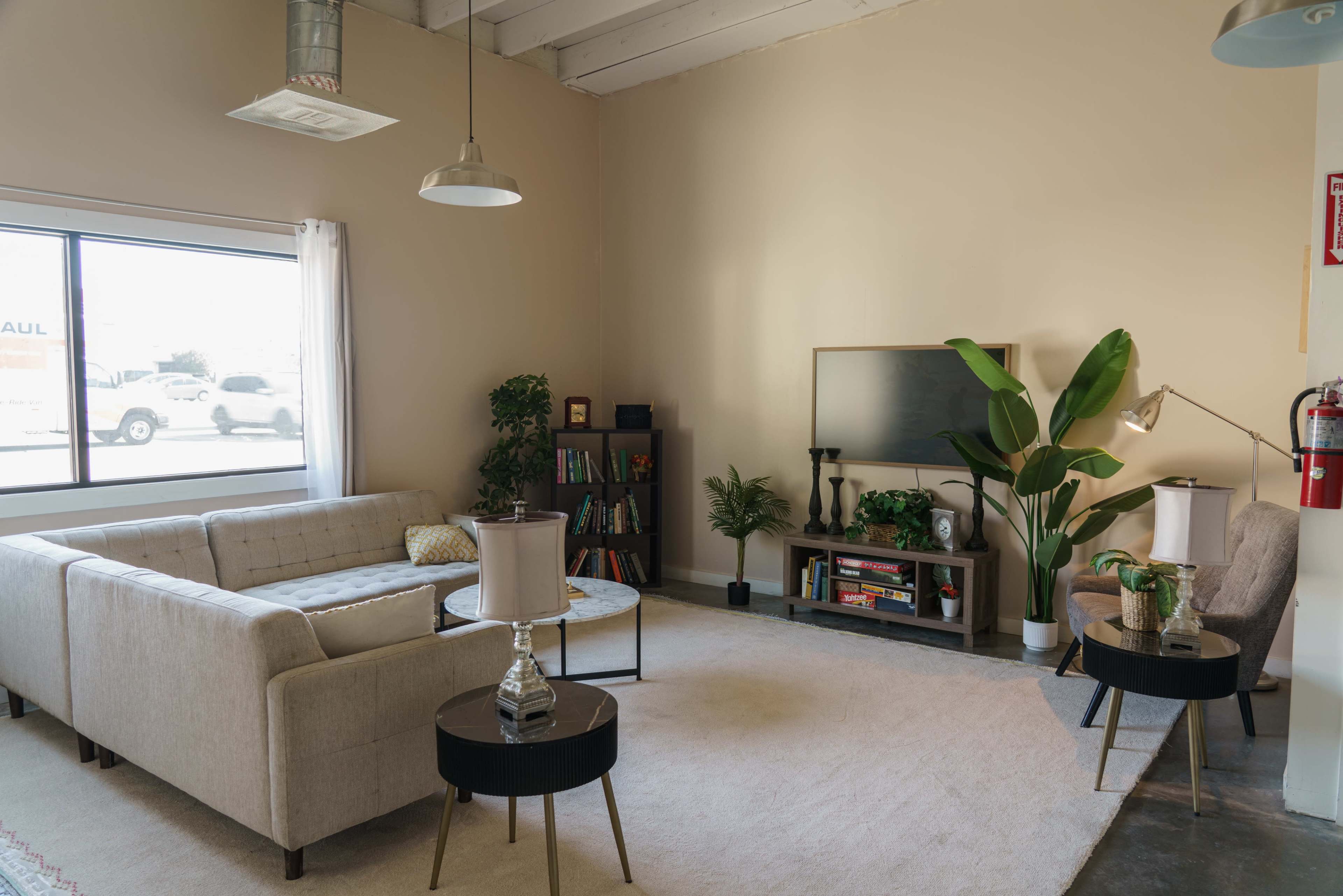 The scene features a modern, minimalist living room with a light beige wall, a gray sofa, two small tables, a bookshelf, a TV on a stand, and a large green plant by the window.