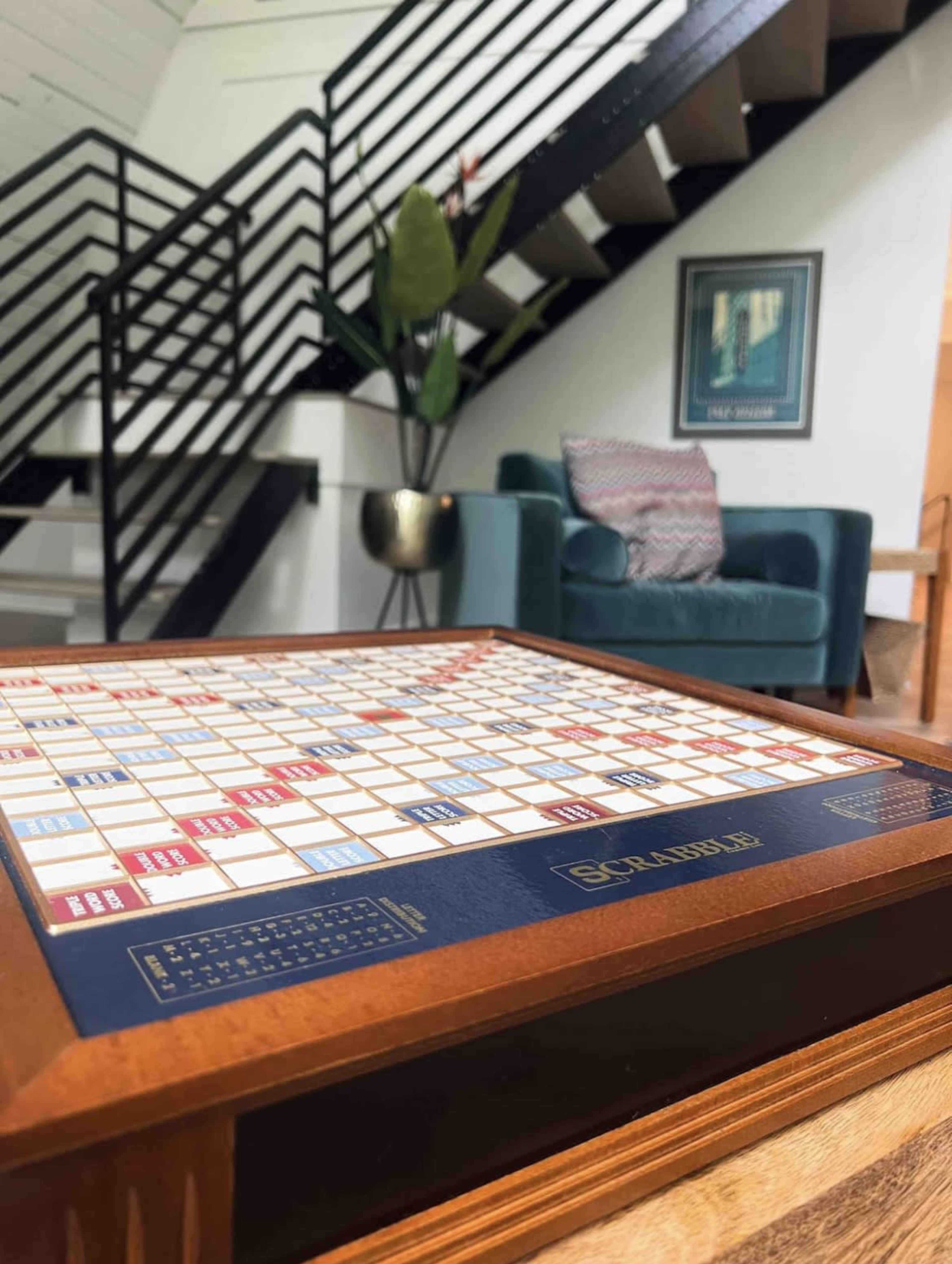 A Scrabble board sits on a wooden table in a living area with a green couch and a staircase in the background.