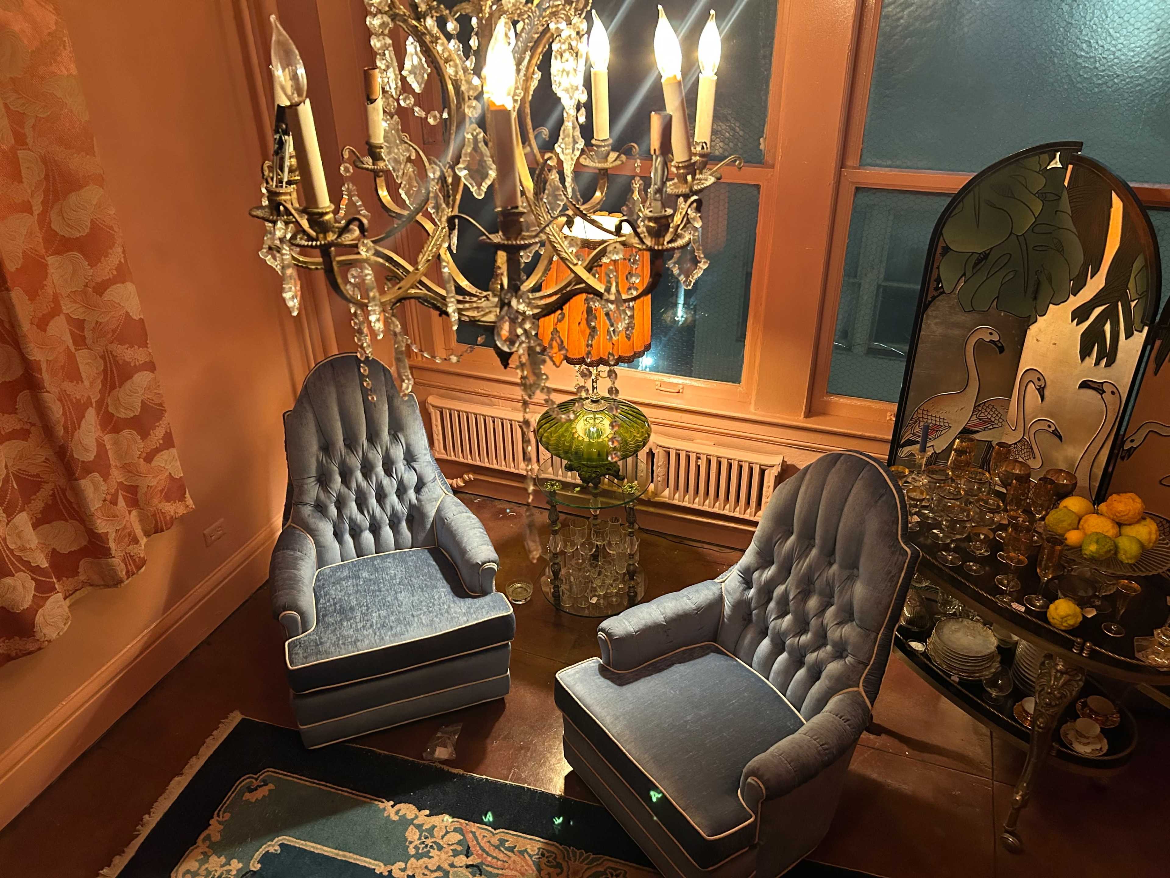 The image shows two blue upholstered chairs positioned near a decorative chandelier and a table with a glass centerpiece and assorted items.