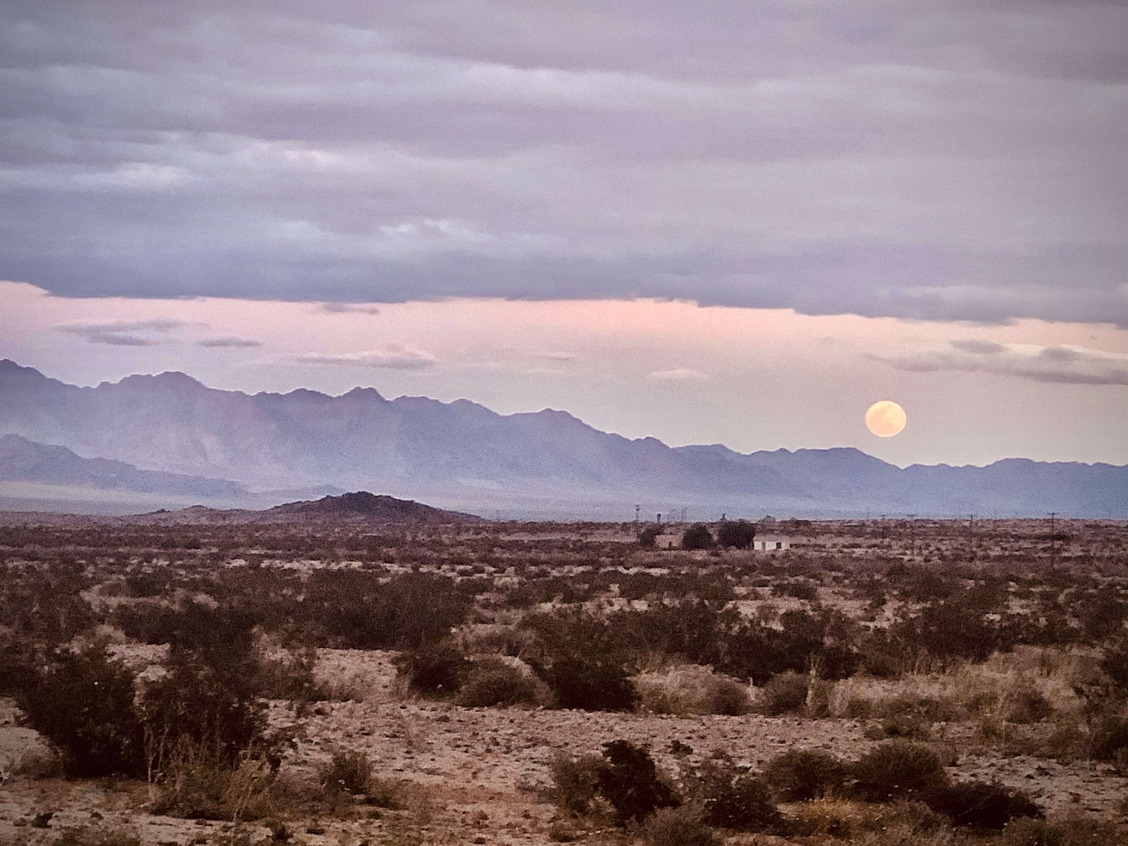A full moon rises over a mountain range in a desert landscape under a cloudy sky.