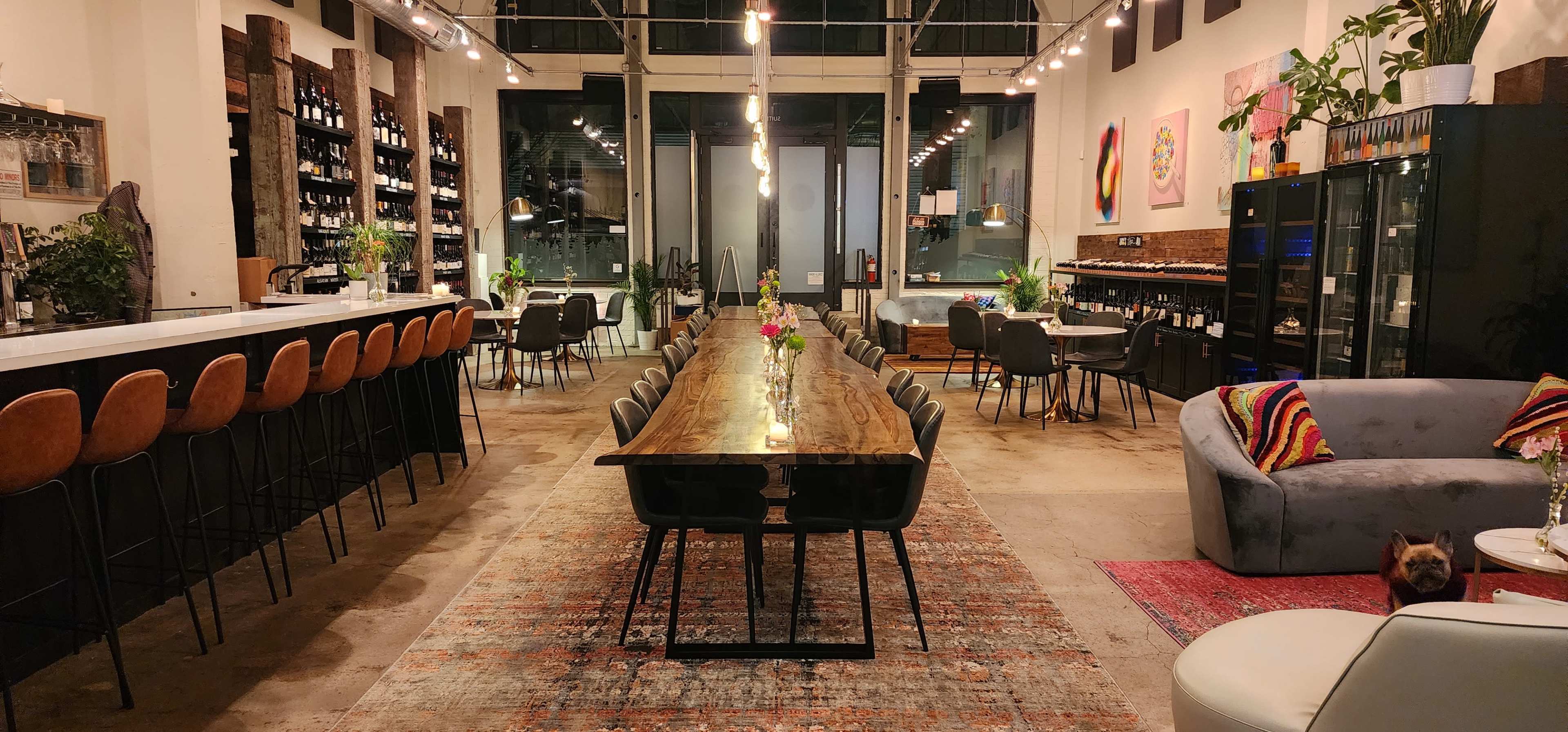 The image shows a spacious, modern interior of a restaurant featuring a long wooden dining table, bar seating, and various seating arrangements with decor and plants.