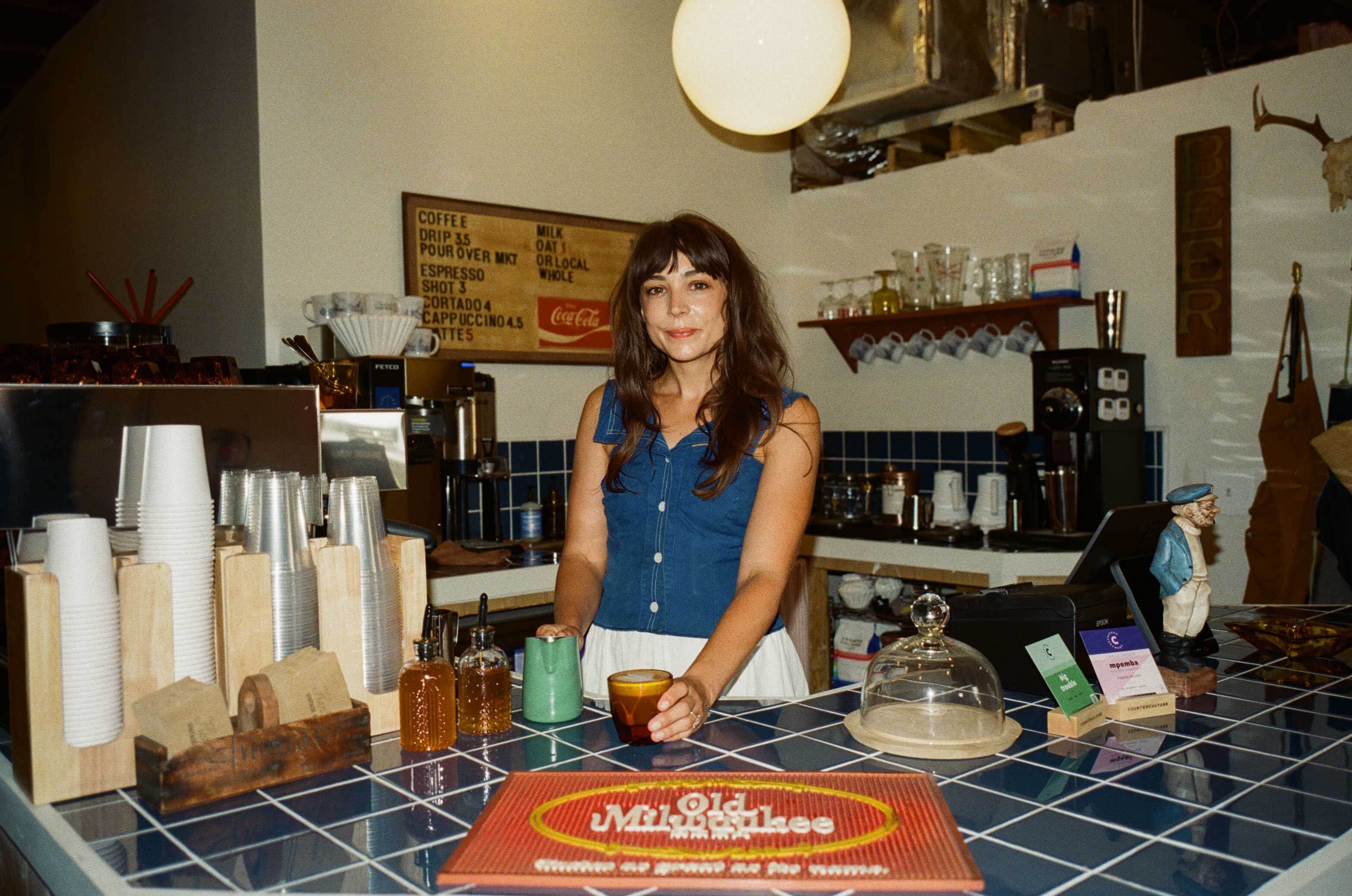 A woman stands at a coffee shop counter, holding a cup and smiling, with various food and drink items behind her.