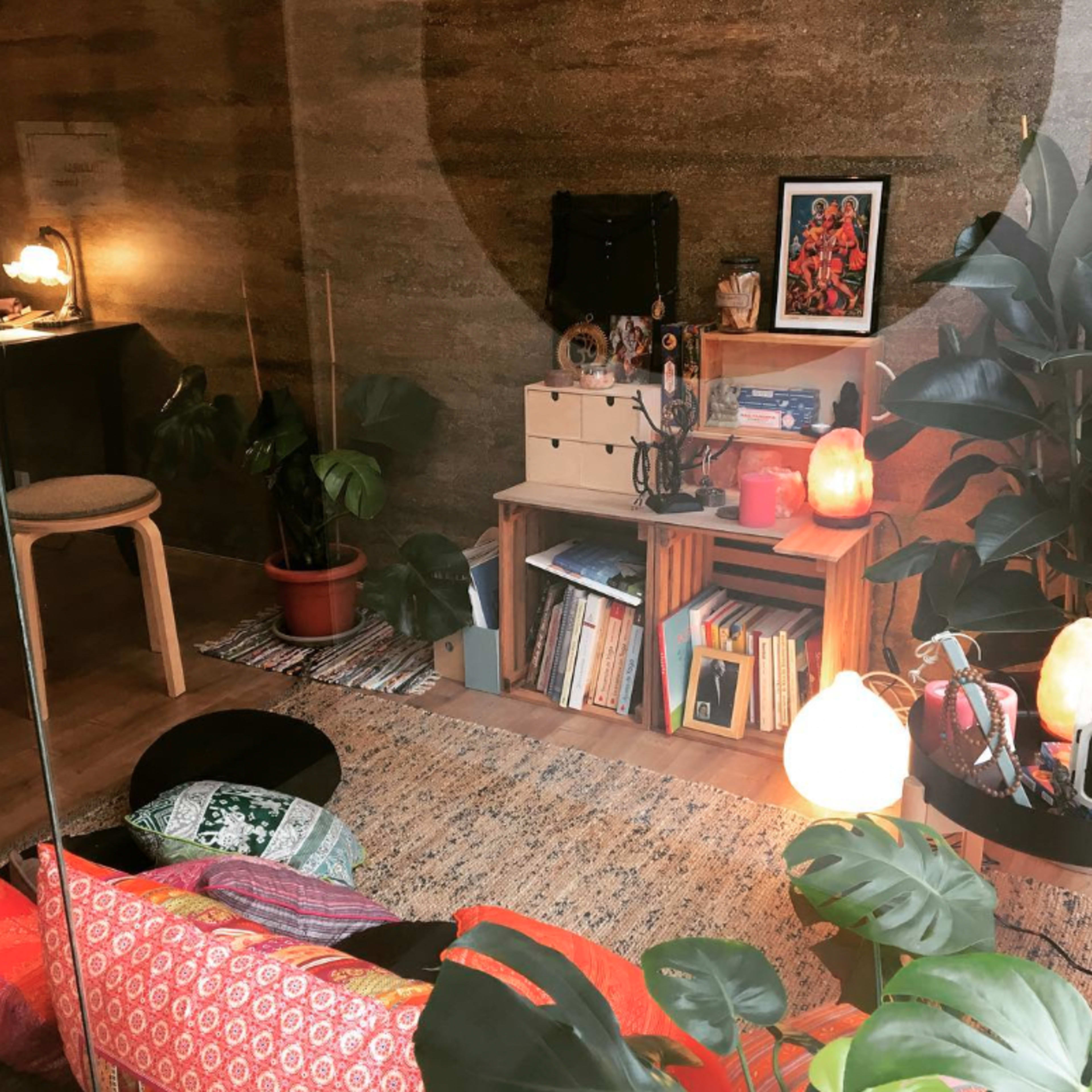 A cozy interior space with a mix of plants, bookshelves, and soft lighting arranged around a seating area on a textured rug.