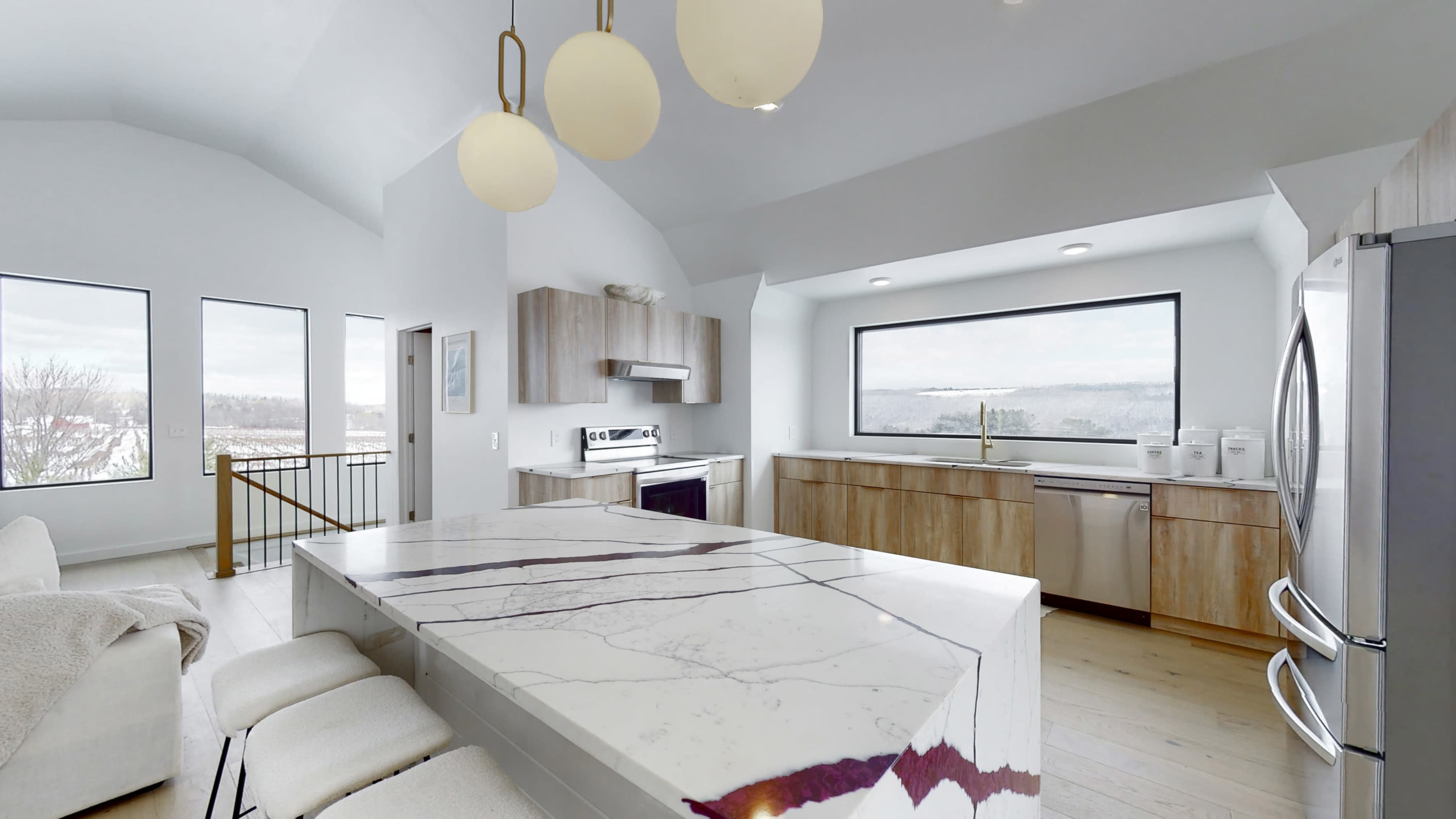 A modern kitchen features a large island with a white and purple marble countertop, wooden cabinetry, and large windows offering a scenic view.