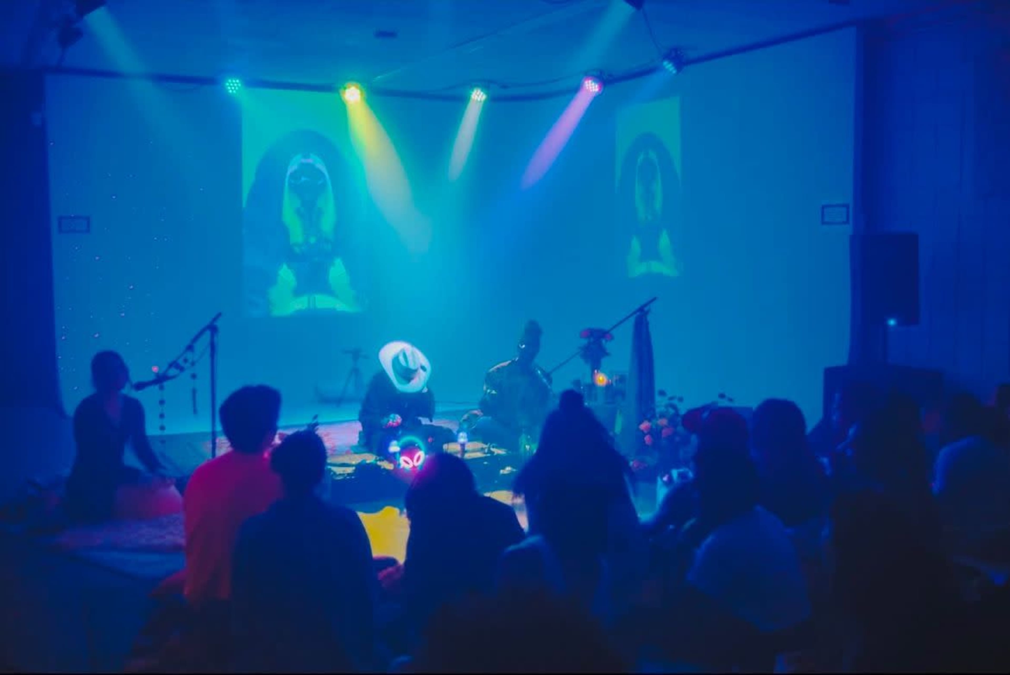 A darkened room filled with an audience seated on the floor, watching performers illuminated by colorful stage lights, with images projected on screens behind them.