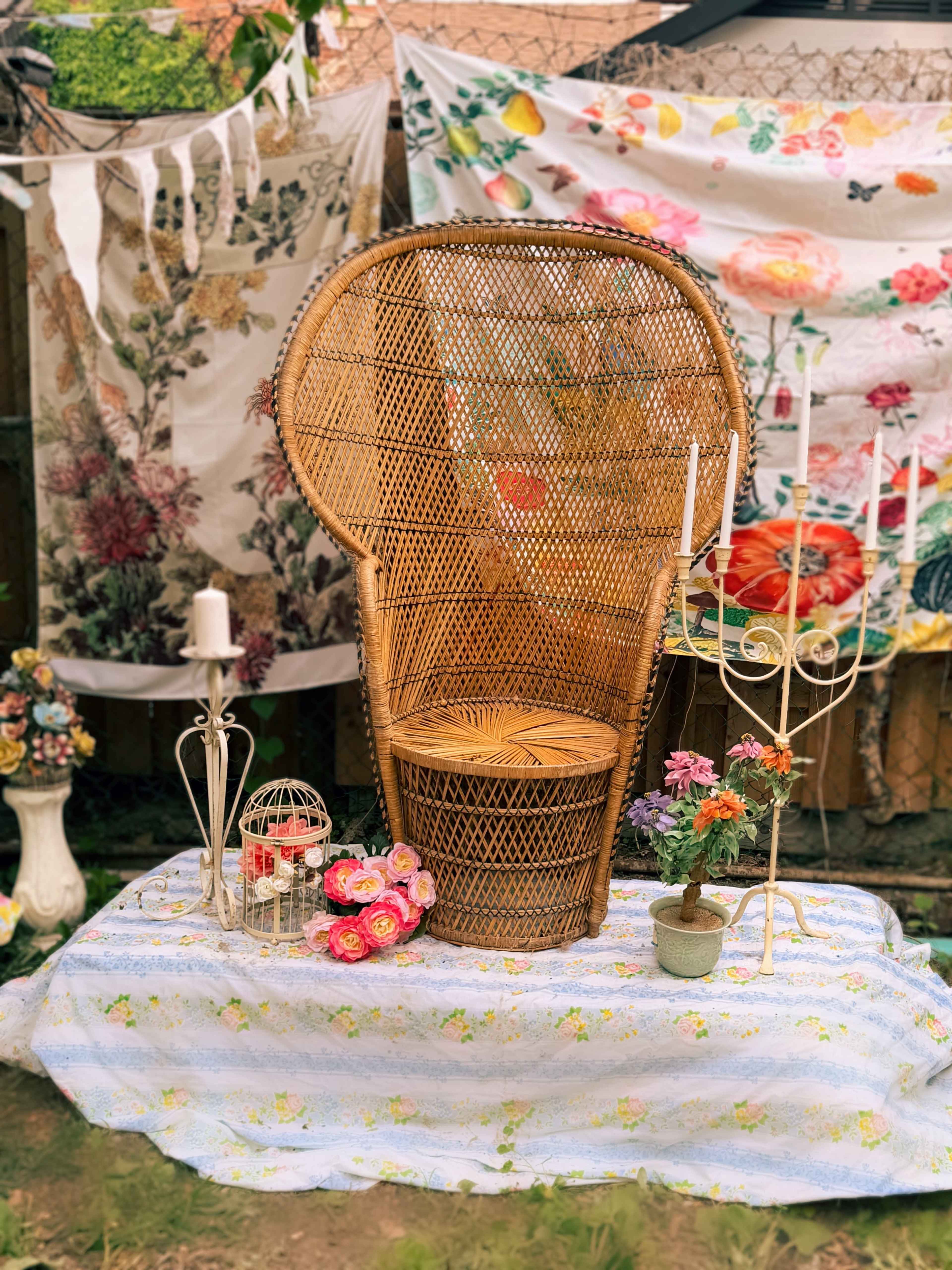A large rattan peacock chair is placed in front of floral-patterned fabrics and surrounded by decorative elements like candles, a birdcage, and flower arrangements on a tablecloth.