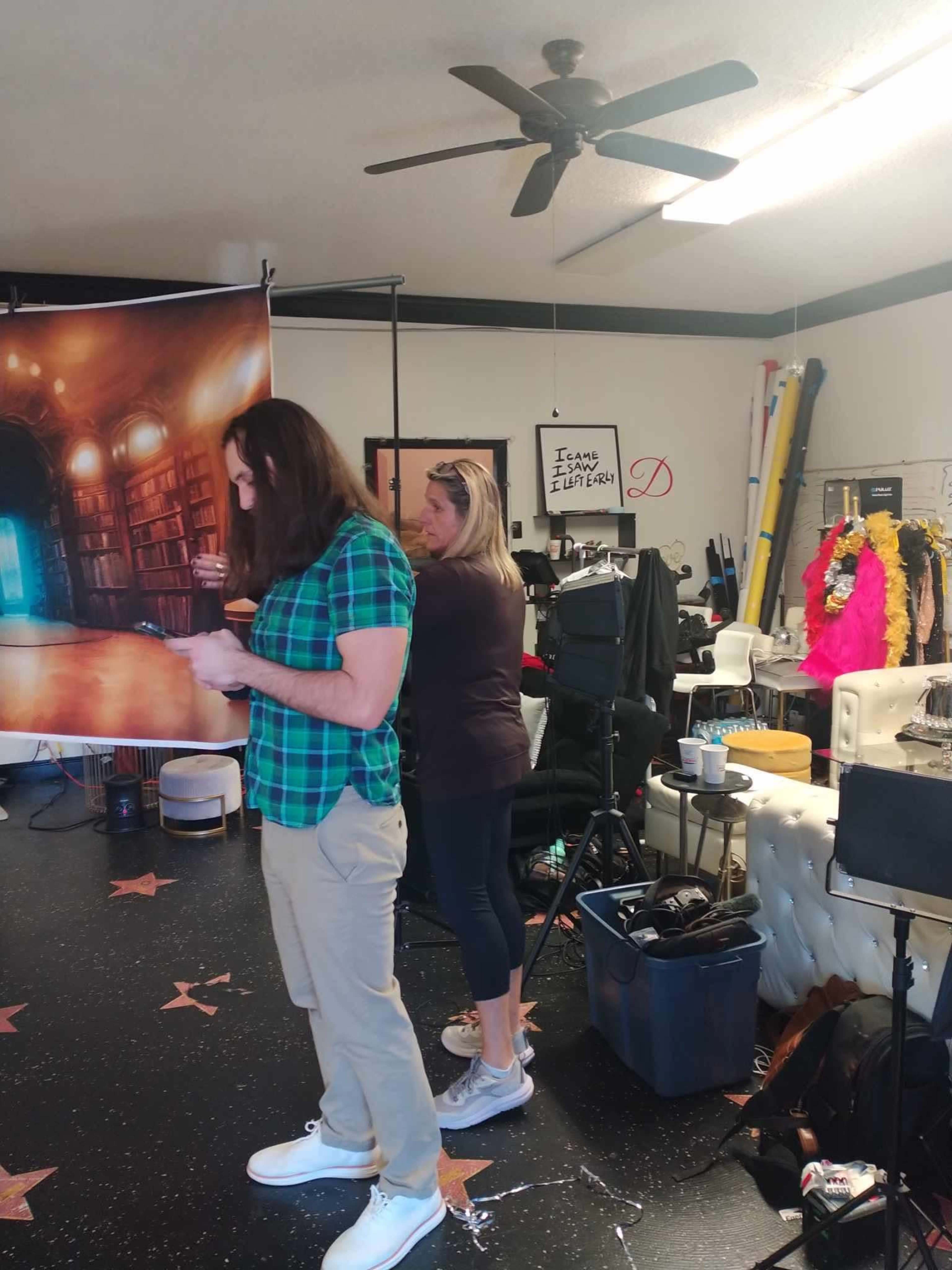 A man in a green checked shirt and a woman in a dark top stand in a room filled with photography equipment and backdrops.