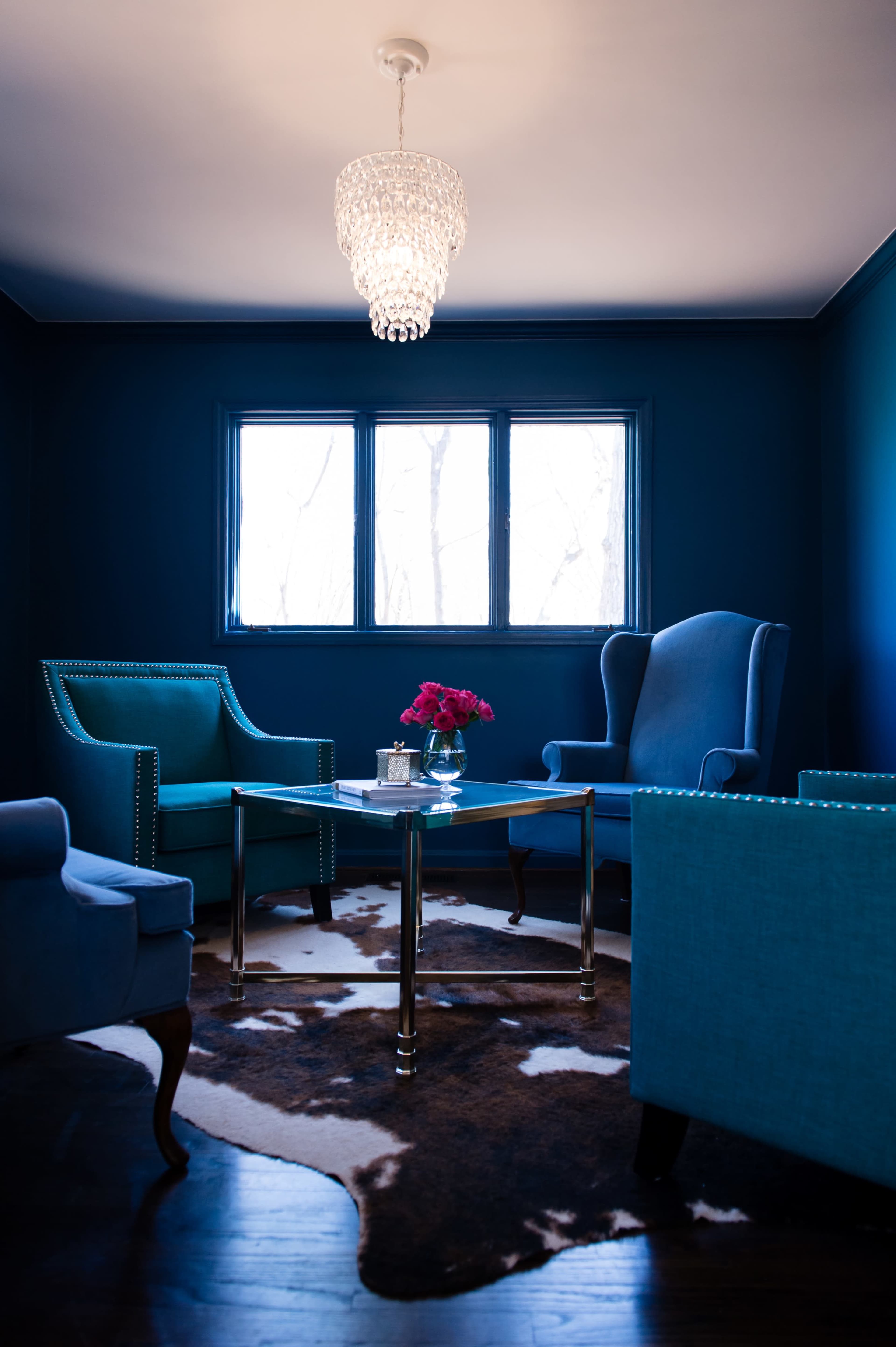 A cozy sitting area features two blue chairs, a glass table with a bouquet of flowers, and a chandelier, all set against a dark blue wall.