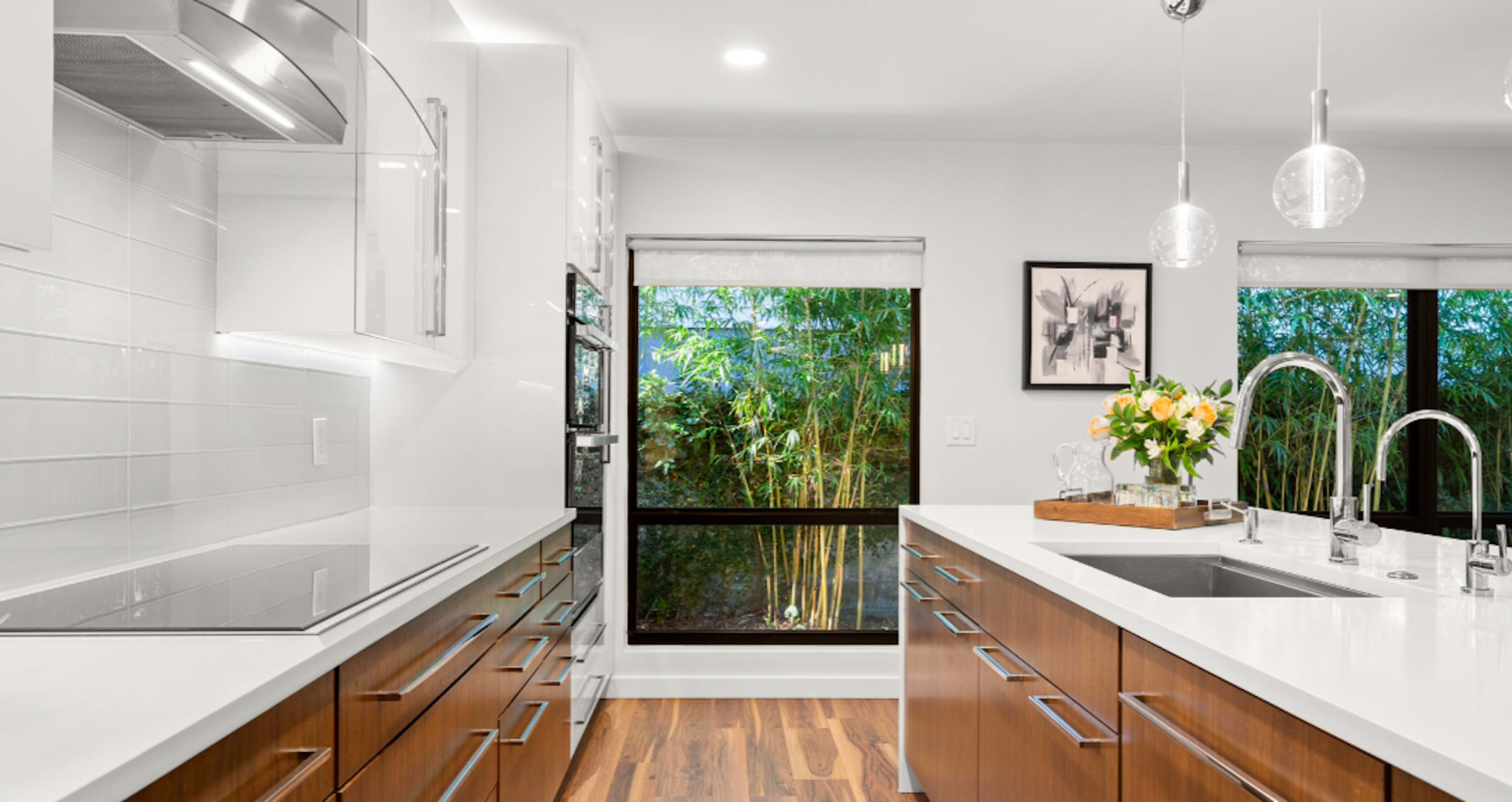 A modern kitchen features sleek cabinetry, a central island with a sink, and large windows showcasing a view of green plants outside.