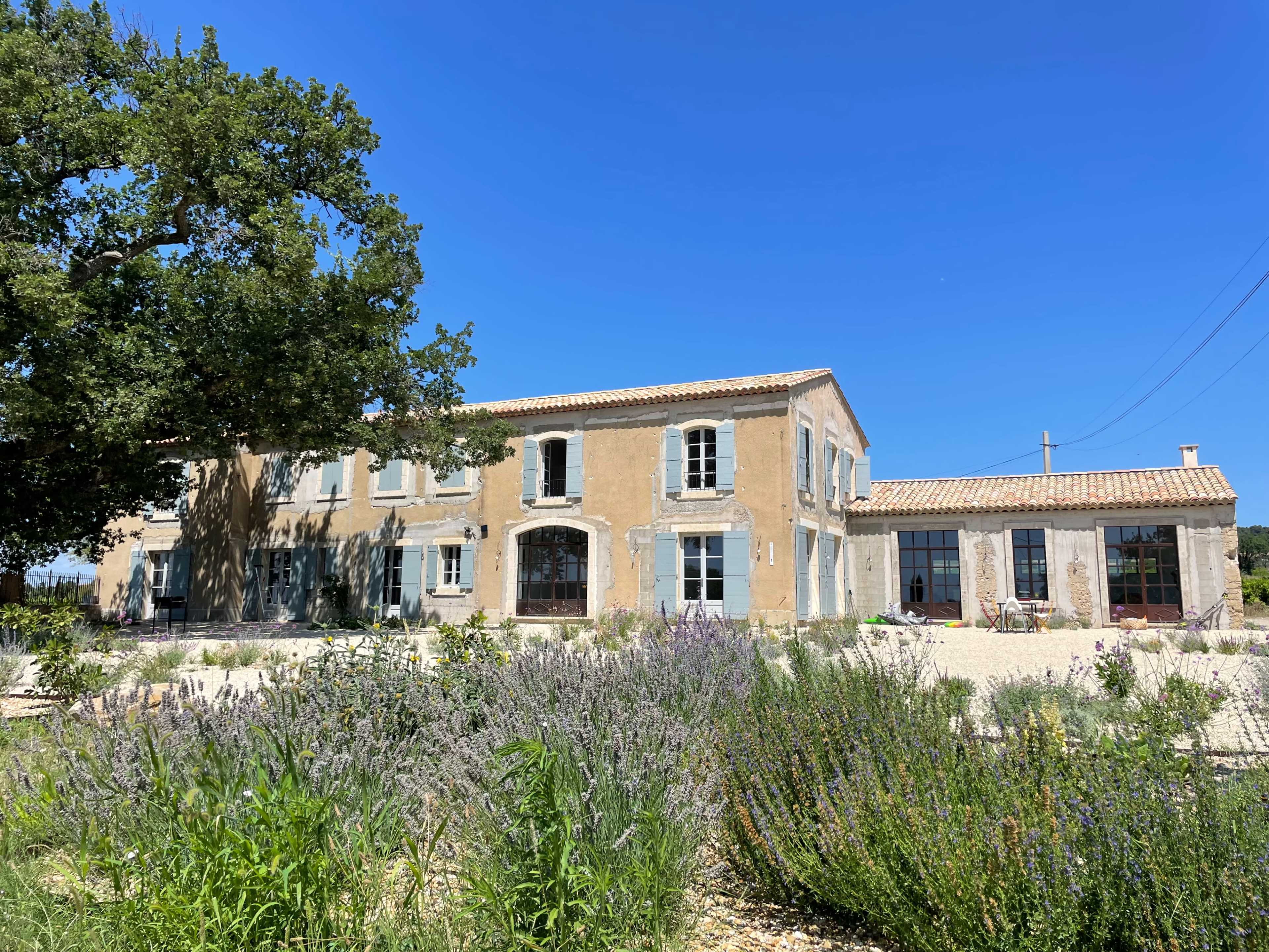 A large, two-story stone house with a tiled roof is set among landscaped gardens featuring lavender and other greenery under a clear blue sky.