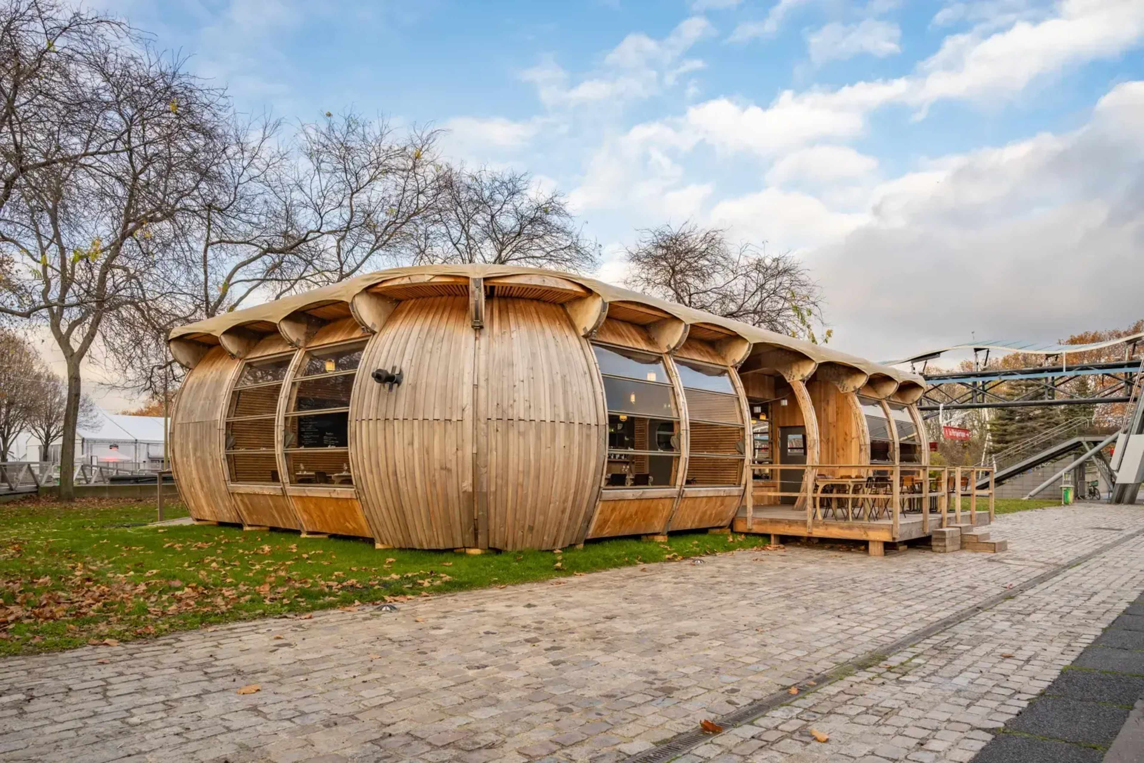 A wooden building with a curved design, featuring large windows and a porch area, situated on a stone pathway surrounded by trees.