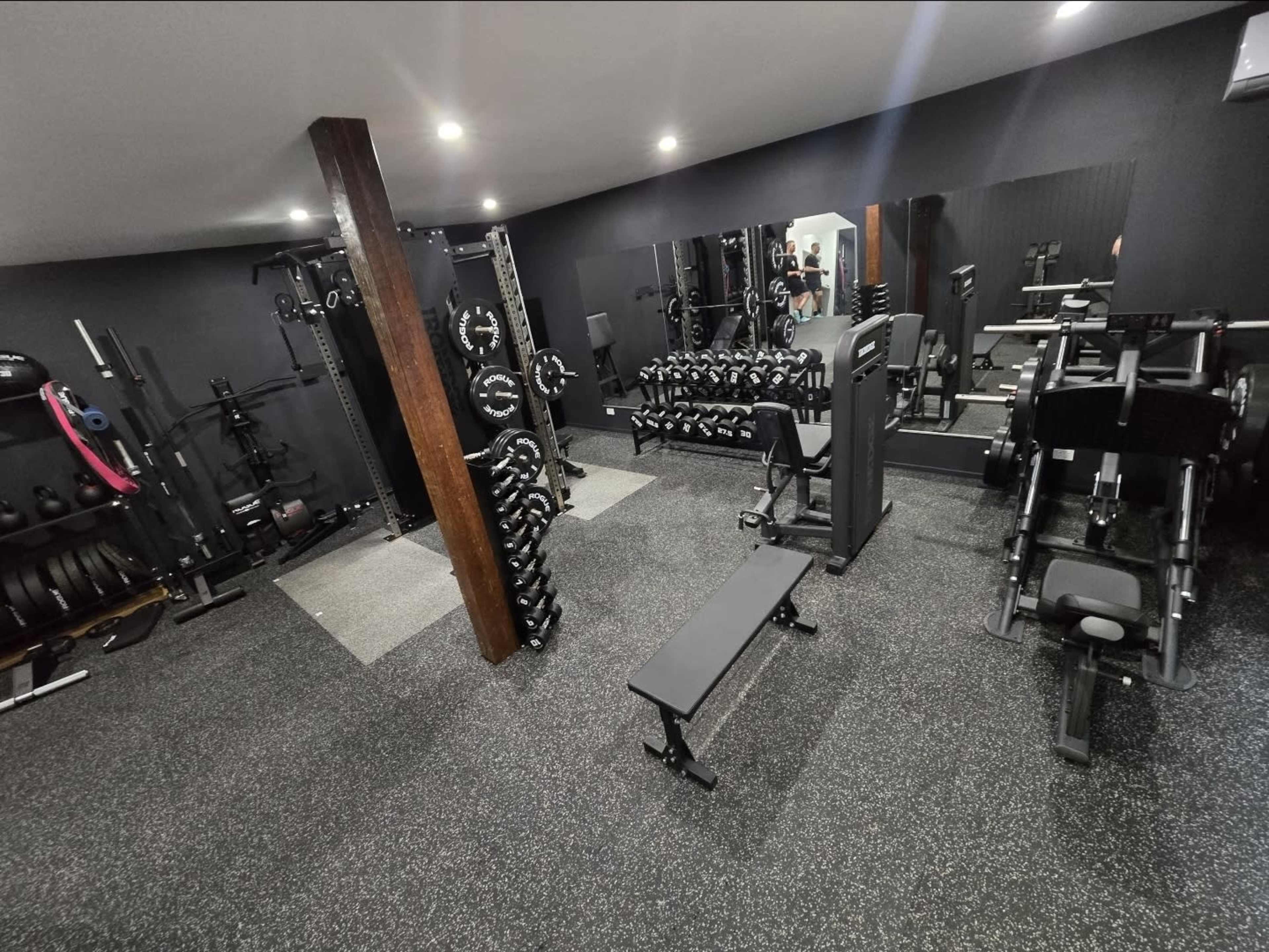 The image shows a well-equipped gym featuring various strength training machines, dumbbells, and free weights on a rubberized floor.