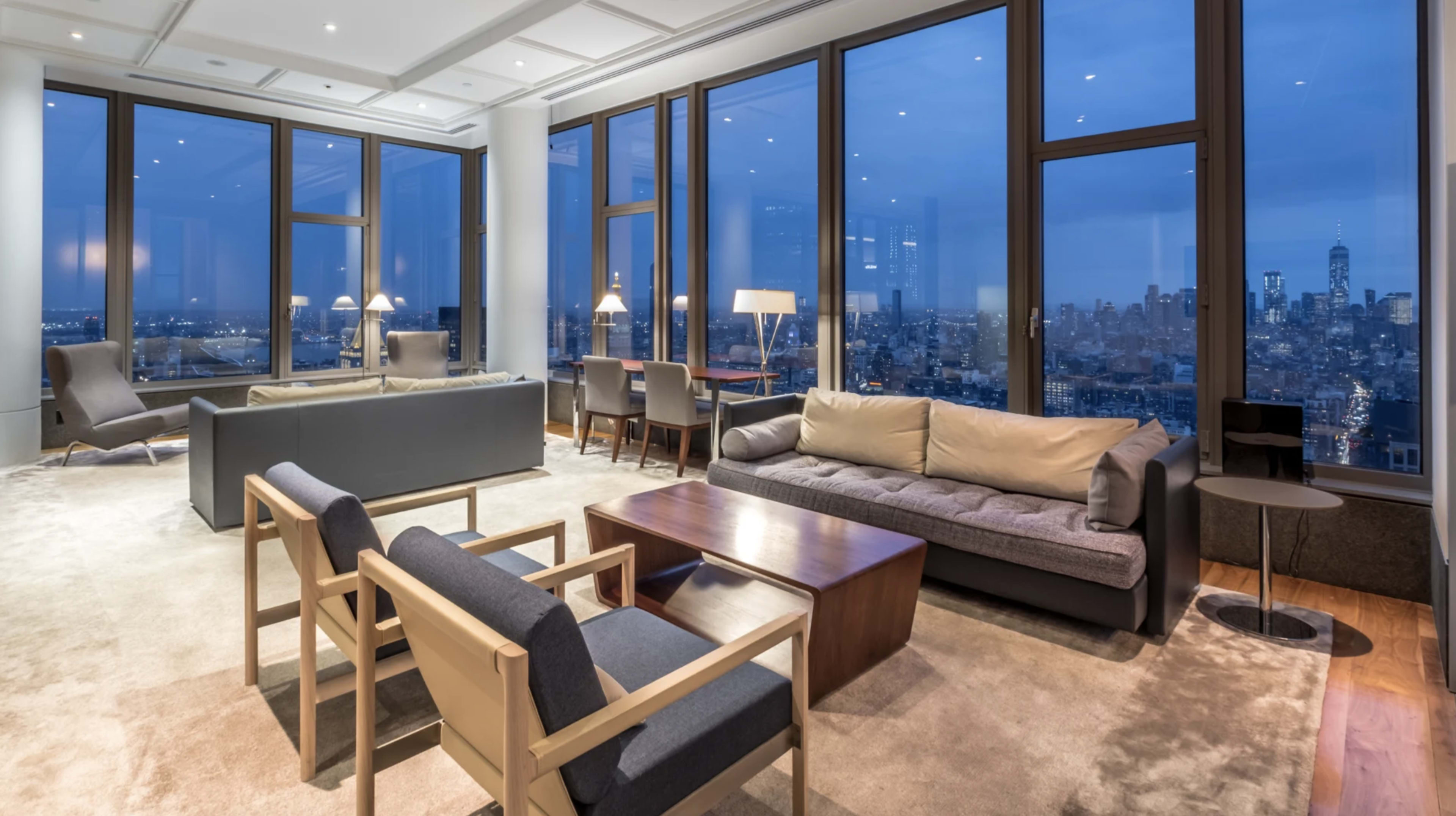 A modern living room with large windows offers a view of a city skyline at dusk, furnished with a sofa, chairs, table, and lamps.