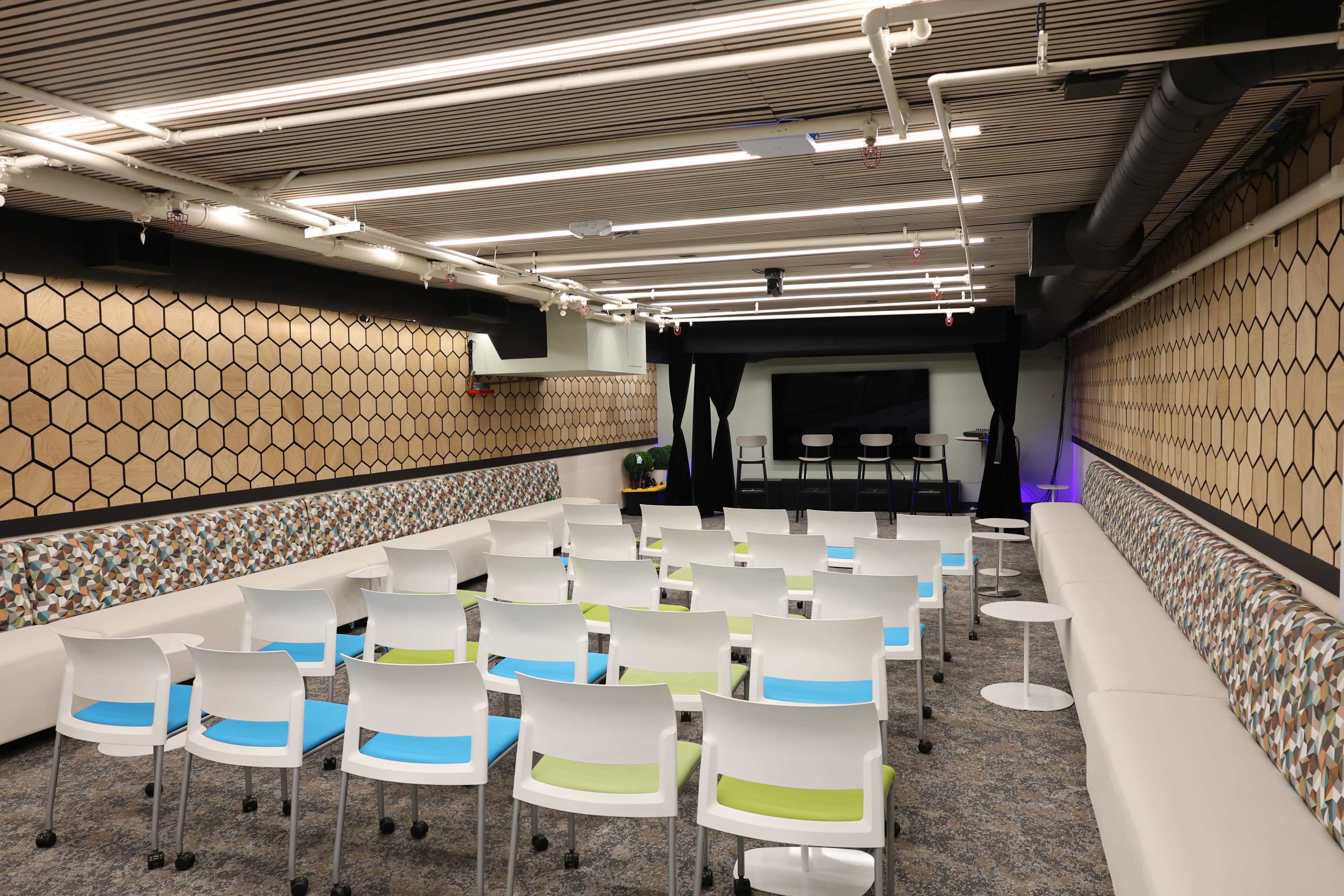 The image shows a modern conference room with hexagonal patterned walls, rows of white chairs with colorful seat cushions, and a stage area with a large screen at the front.