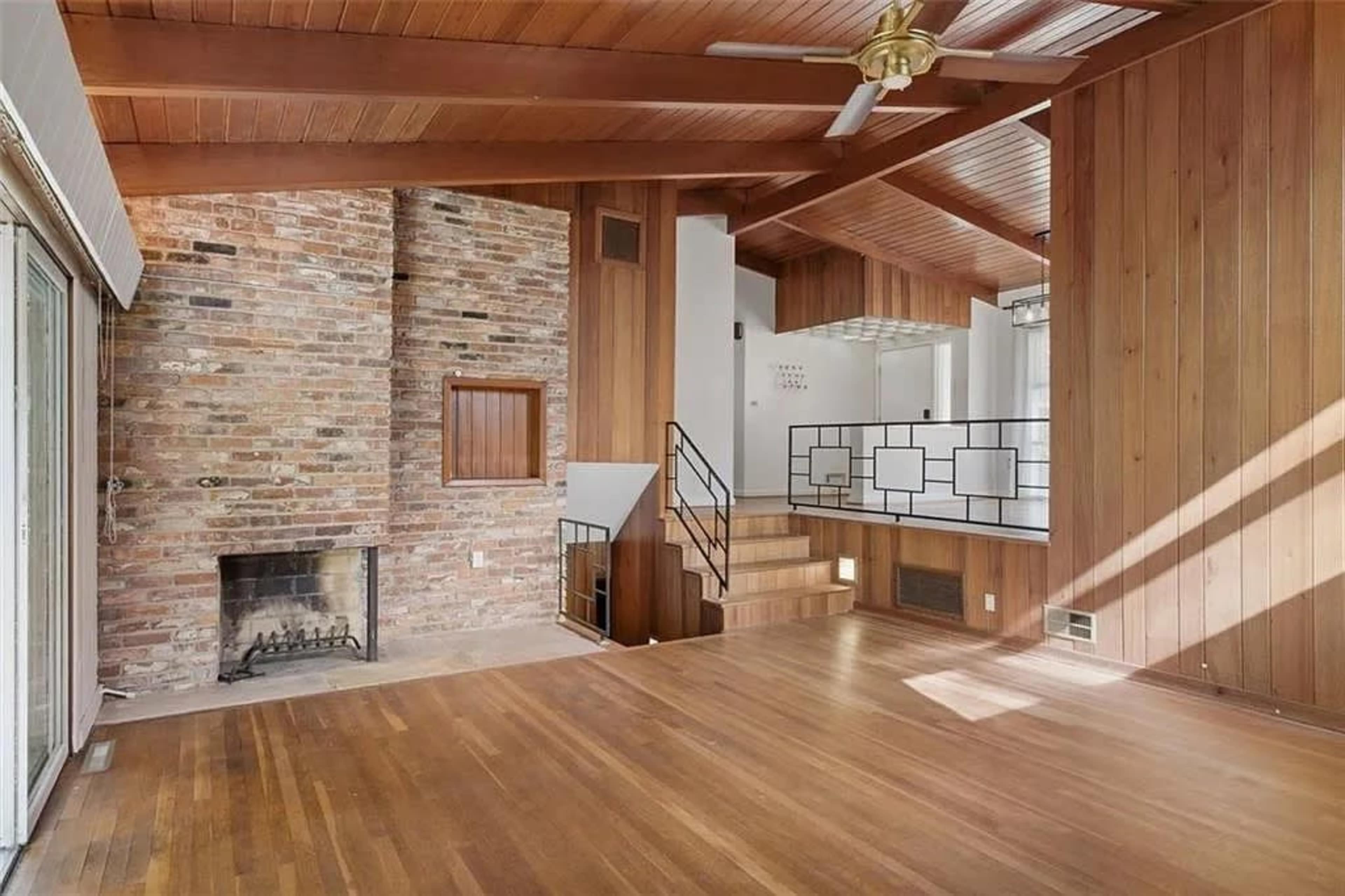 The room features a brick fireplace, wooden walls and ceiling, and a staircase with a railing that leads to an upper level.