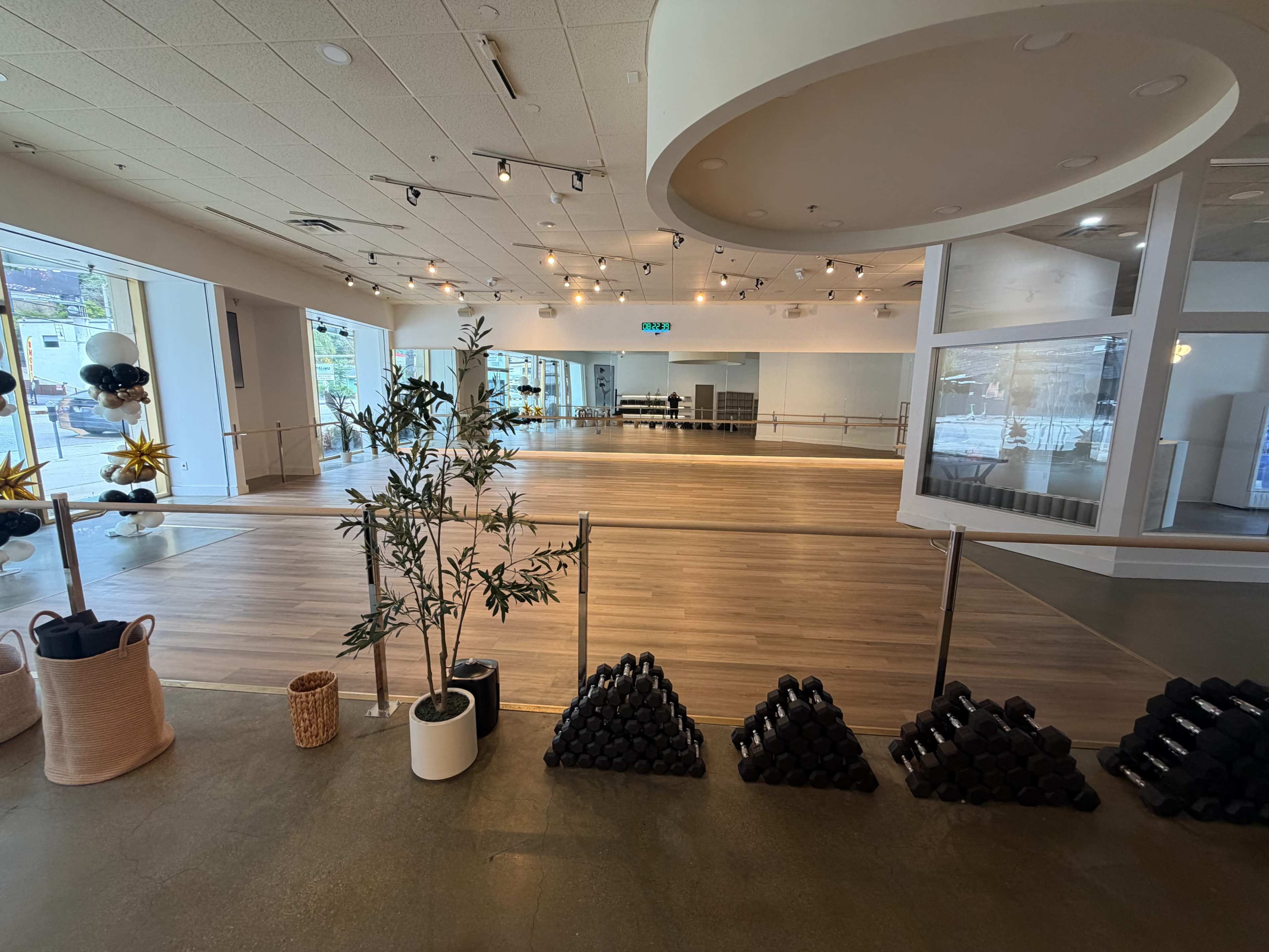 The image shows a spacious fitness studio with wooden floors, large mirrors, and a rack of black weights arranged on the floor.
