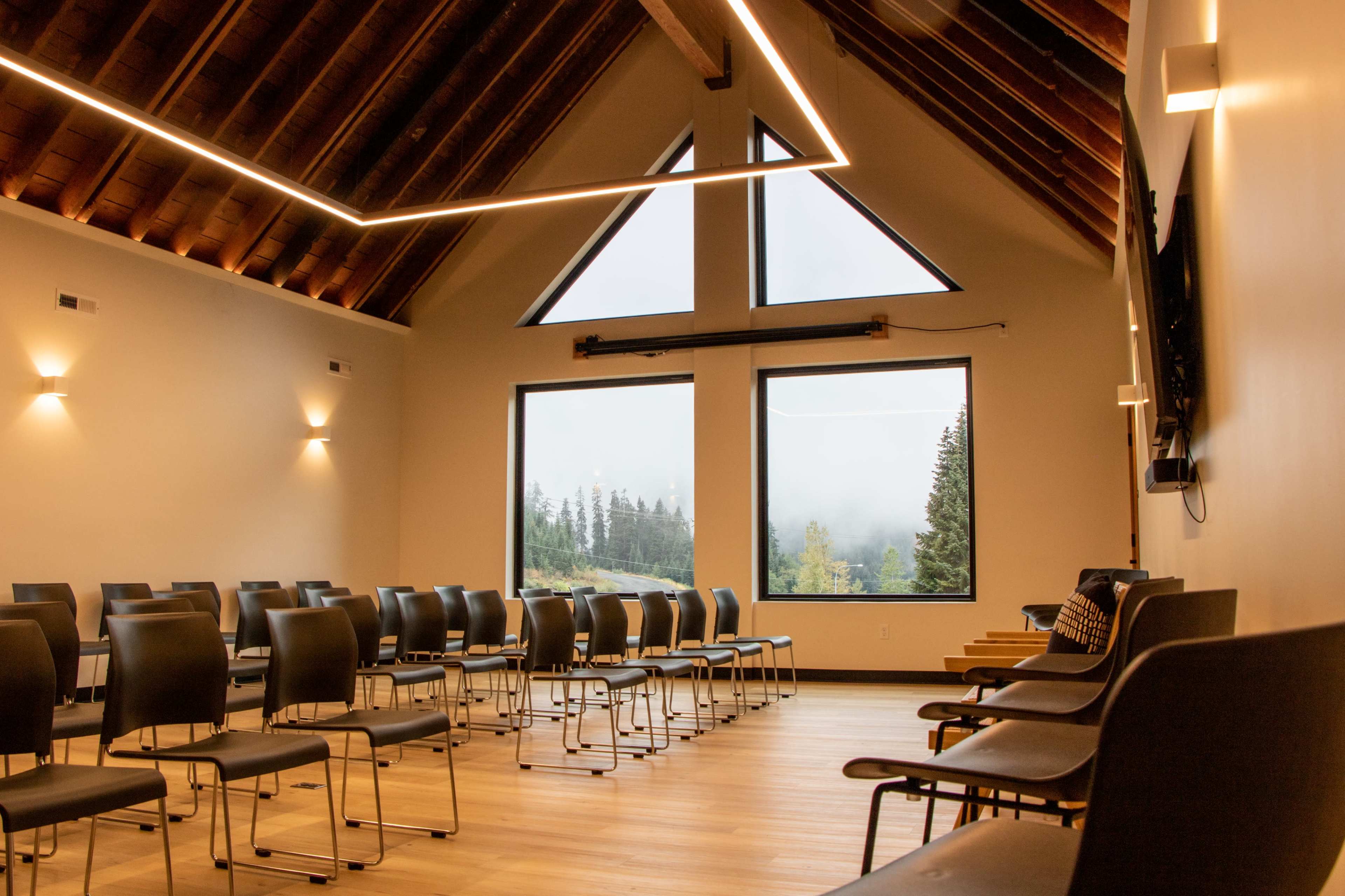 The image shows an empty room with a wooden ceiling, arranged with black chairs facing a large window that overlooks a misty forest.