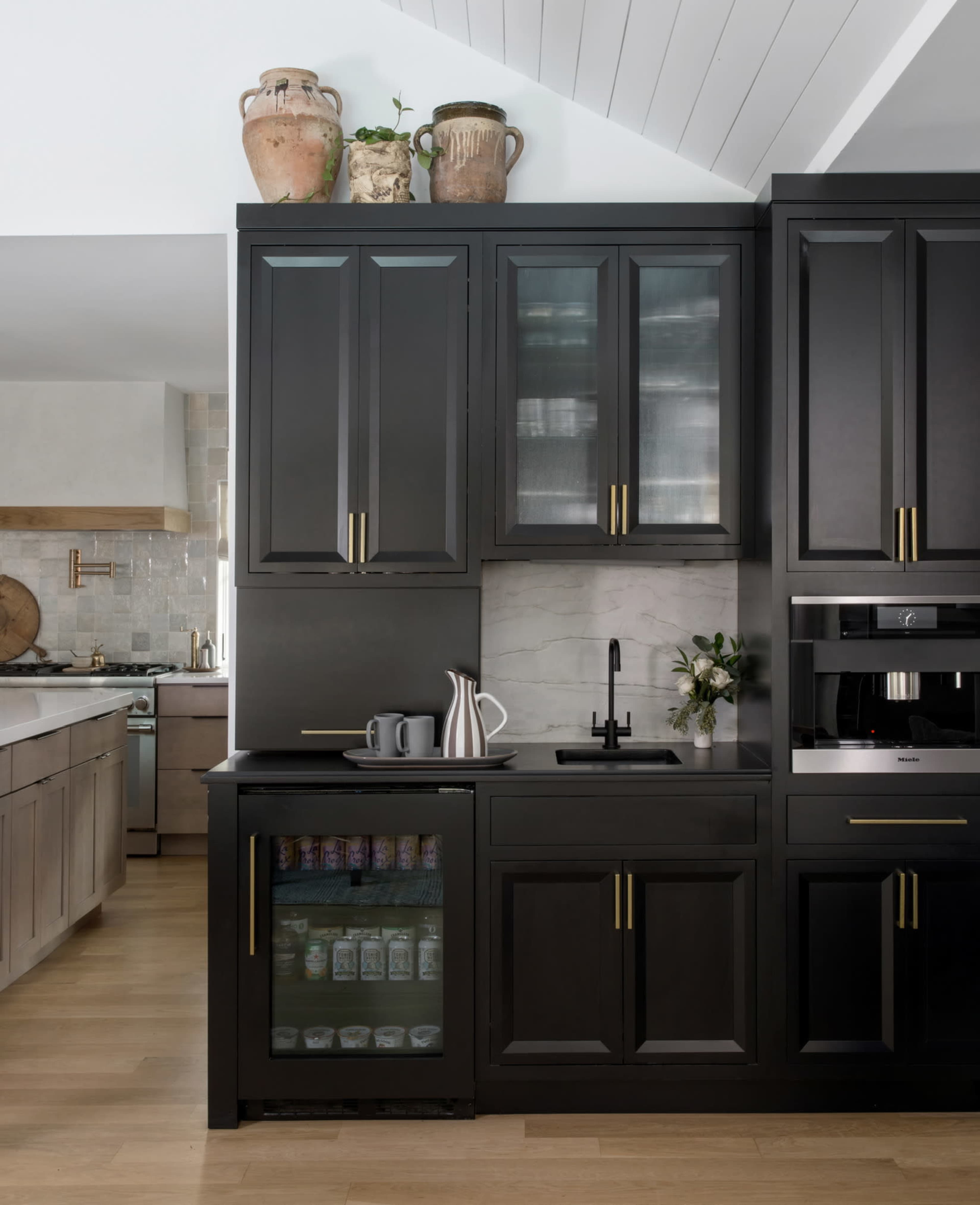 A modern kitchen with dark cabinetry, gold hardware, a small refrigerator with glass doors, and a countertop featuring a sink and decorative items.